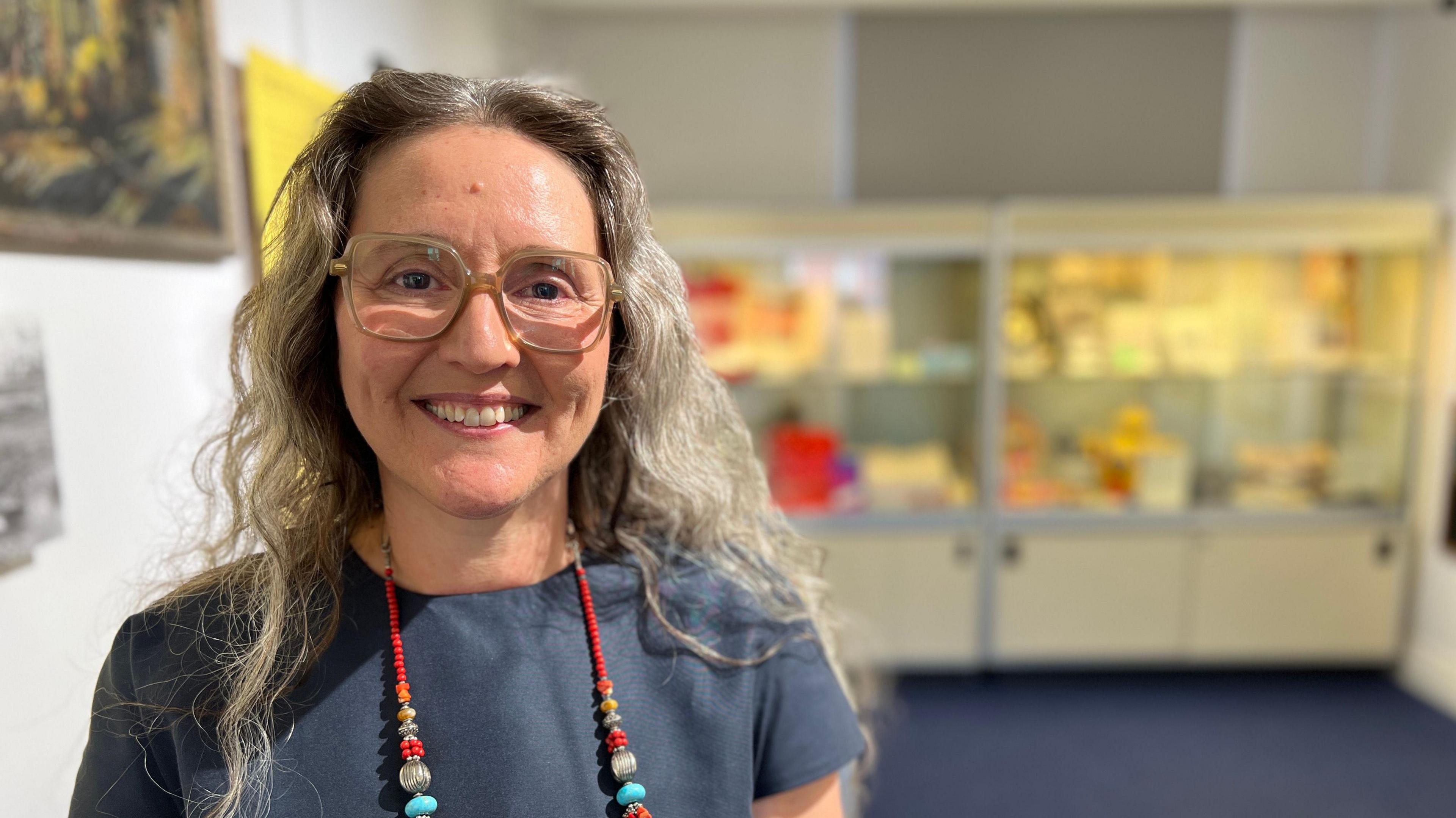 Councillor Marina Strinkovsky, standing in the new gallery. She has long grey, curly hair and wears large-framed glasses and a red beaded necklace. 