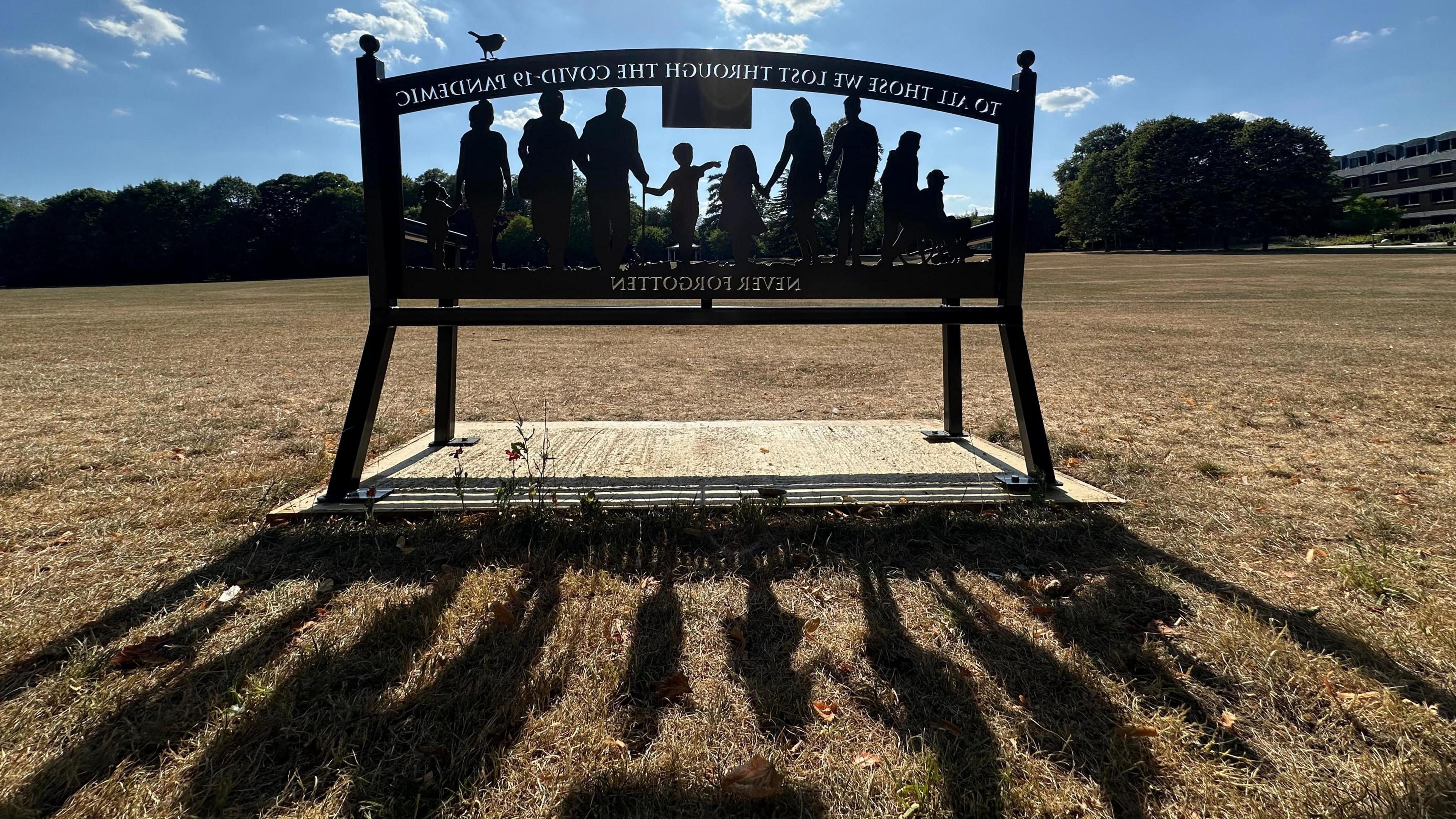 The sun streams through a metal bench made with the silhouetted figures of a group of people, from old to young. The backwards dedication reads: "To all those lost through the Covid-19 pandemic. Never Forgotten."