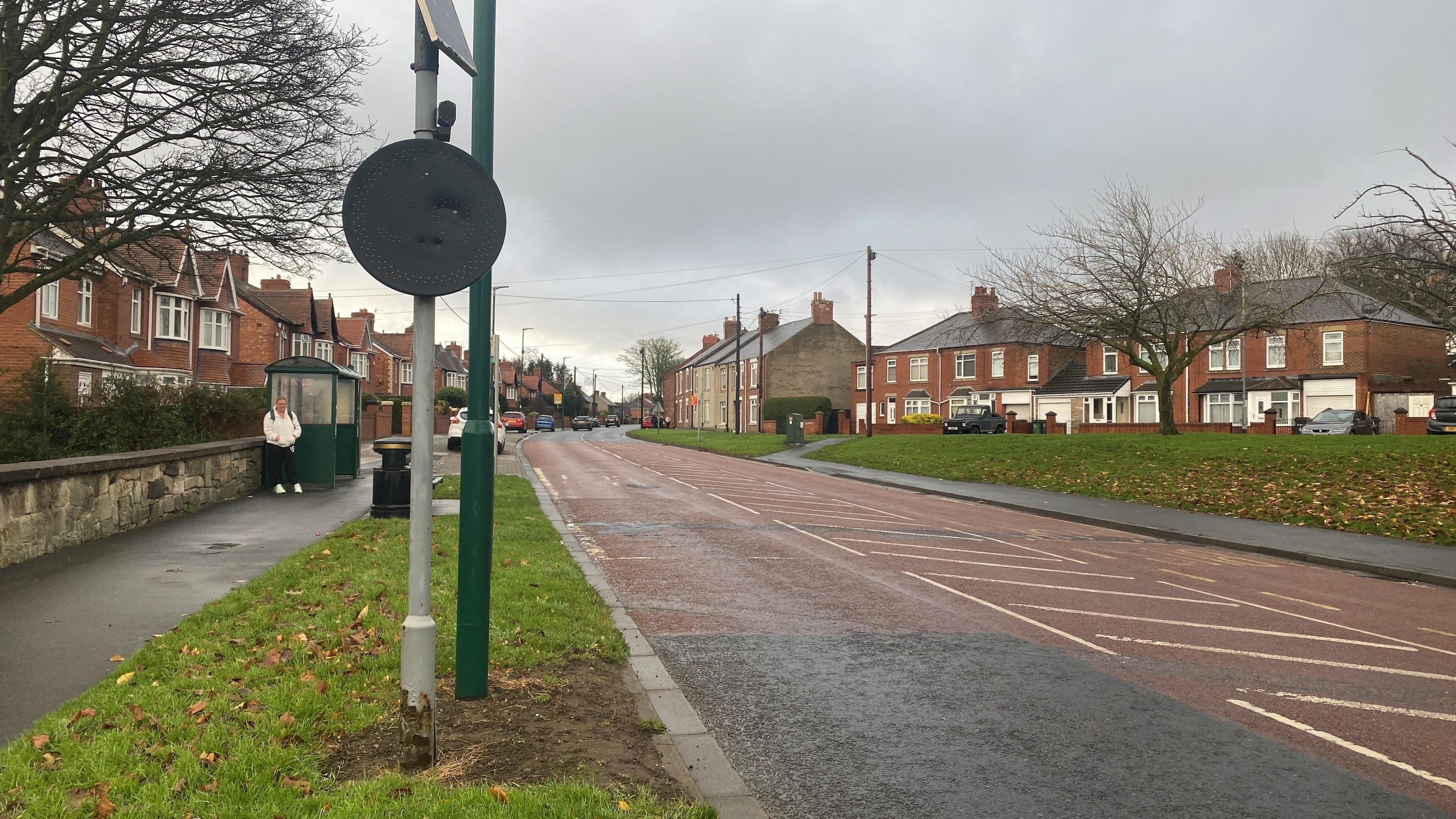 A wide road in Bedlington with 1950s style houses on one side, and older houses on the right. There are wide grass verges.