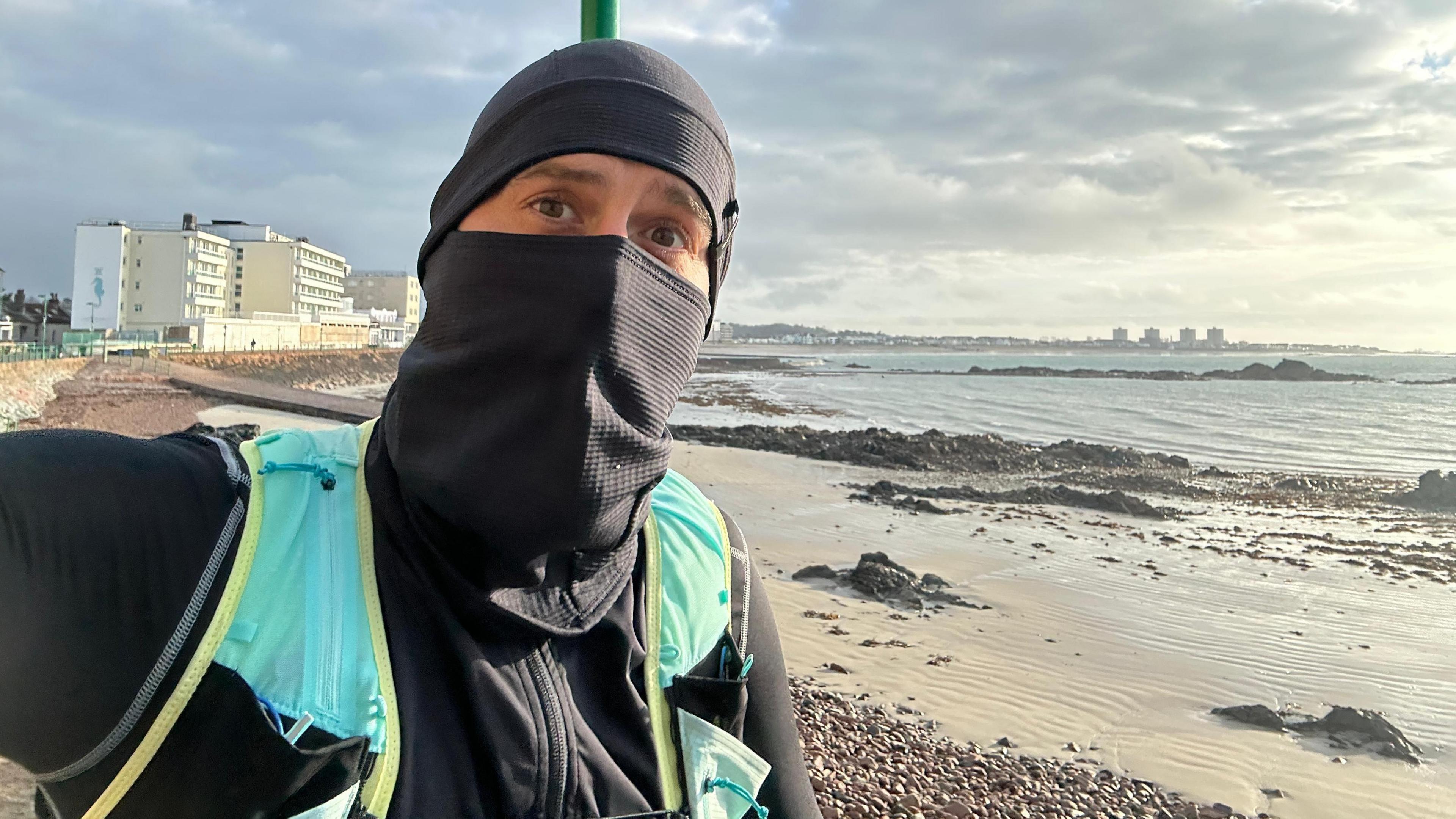 Sam is taking a selfie as he runs along a sea front with a beach behind him. The sun is breaking through some grey clouds. Sam is wearing a black running jumper with a neck scarf pulled over his mouth and a black hat on his head. He's also wearing a light blue running vest as well.