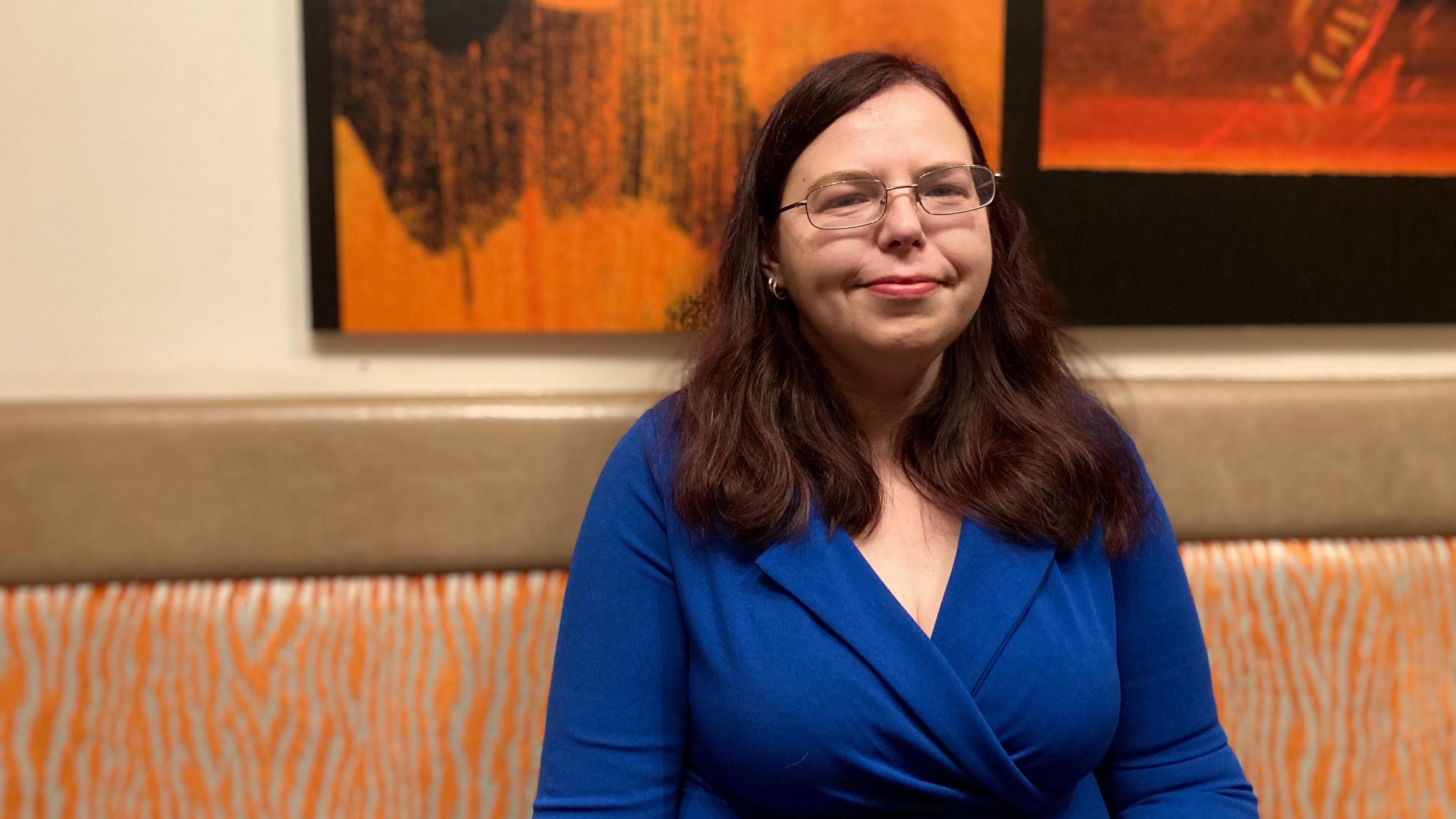 Lucy Robinson smiling into the camera as she sits on a bench in a pub. She has long dark hair, glasses and is wearing a blue top.