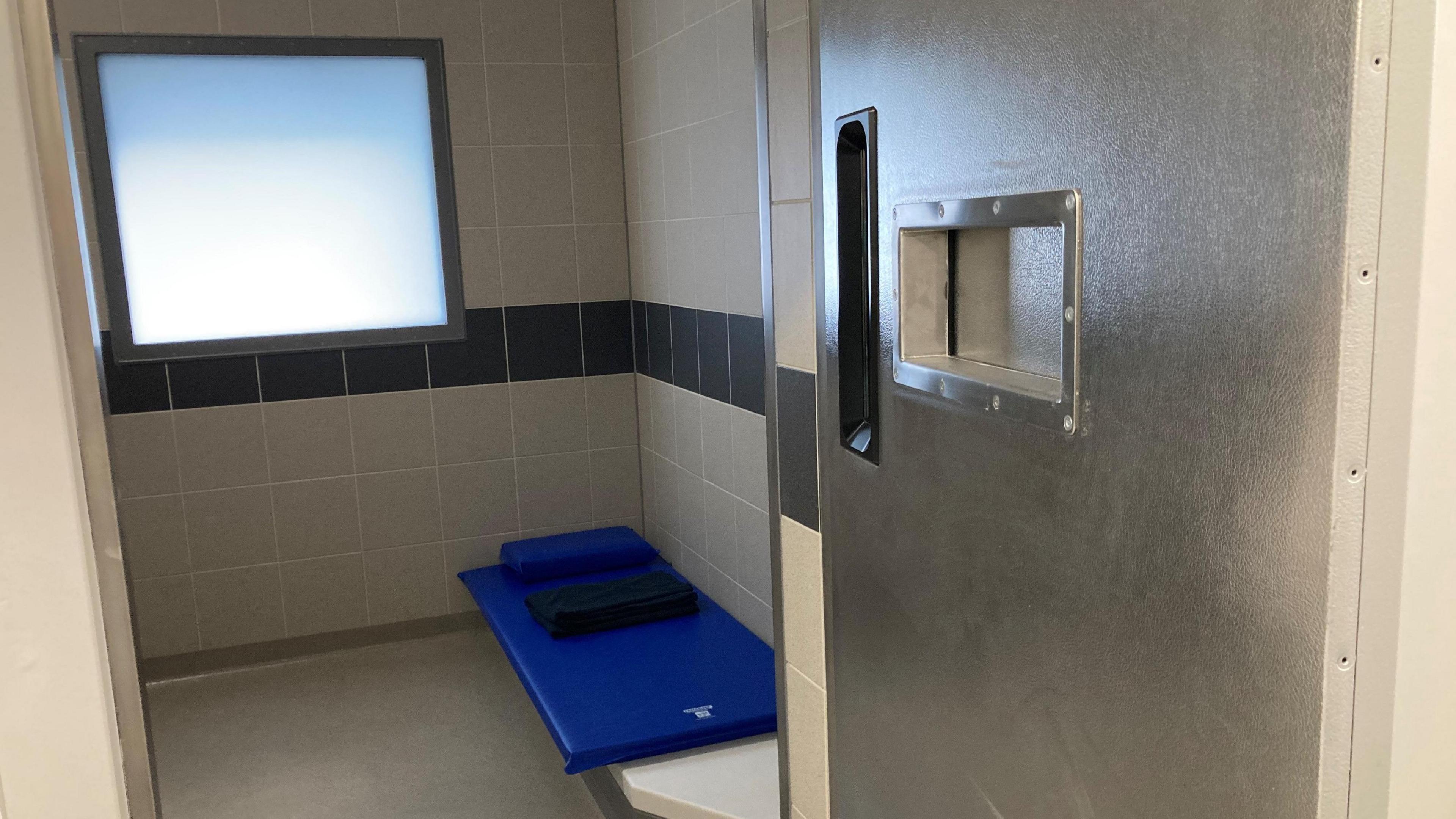 A new police cell. There is a frosted window and tiled walls with a basic blue mattress for a bed.