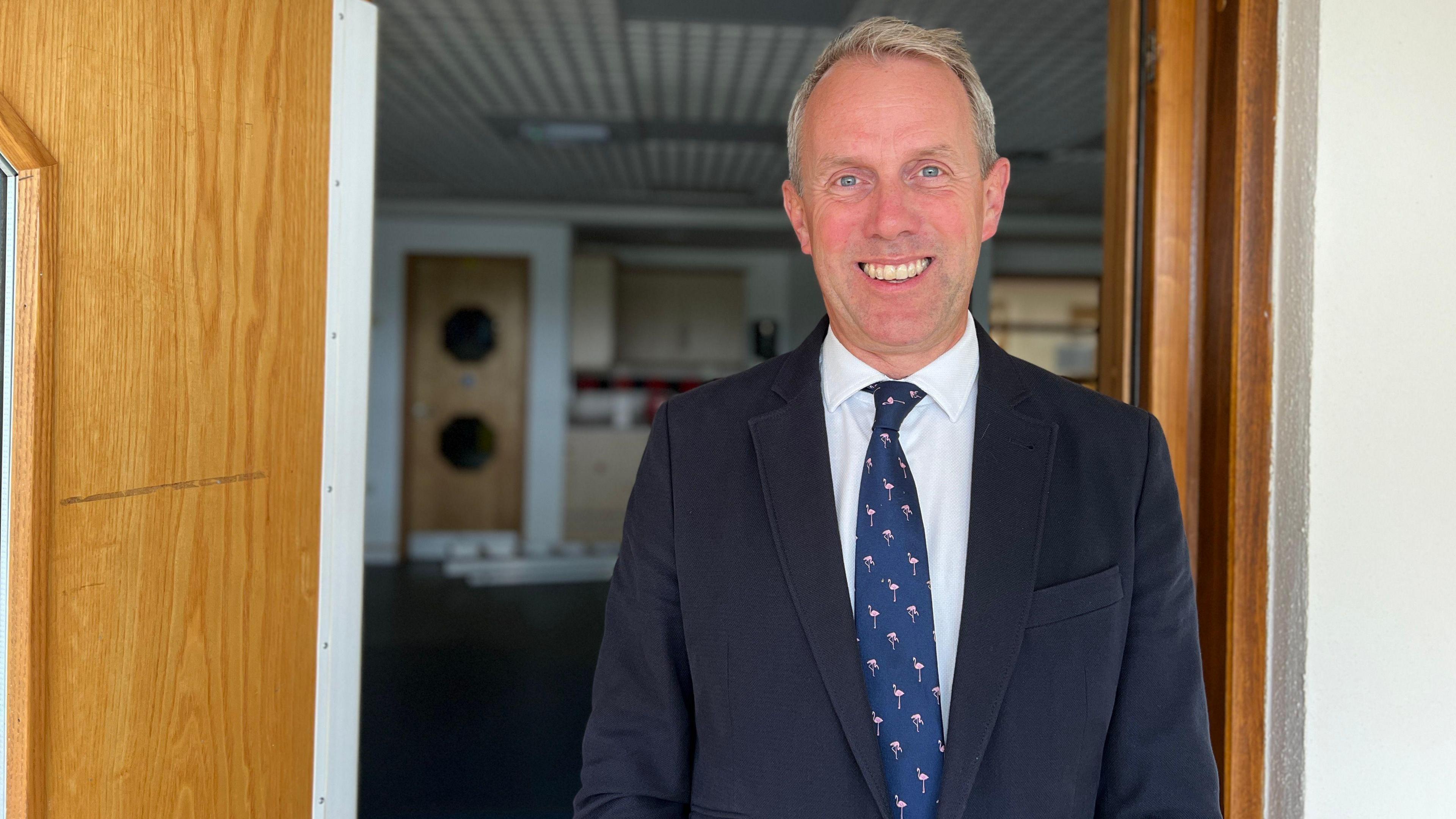 Sam Cooper - man with light hair smiling with teeth, wearing a navy suit, white shirt underneath and a blue tie with mini flamingos on it, standing in the doorway, behind is blurred school room