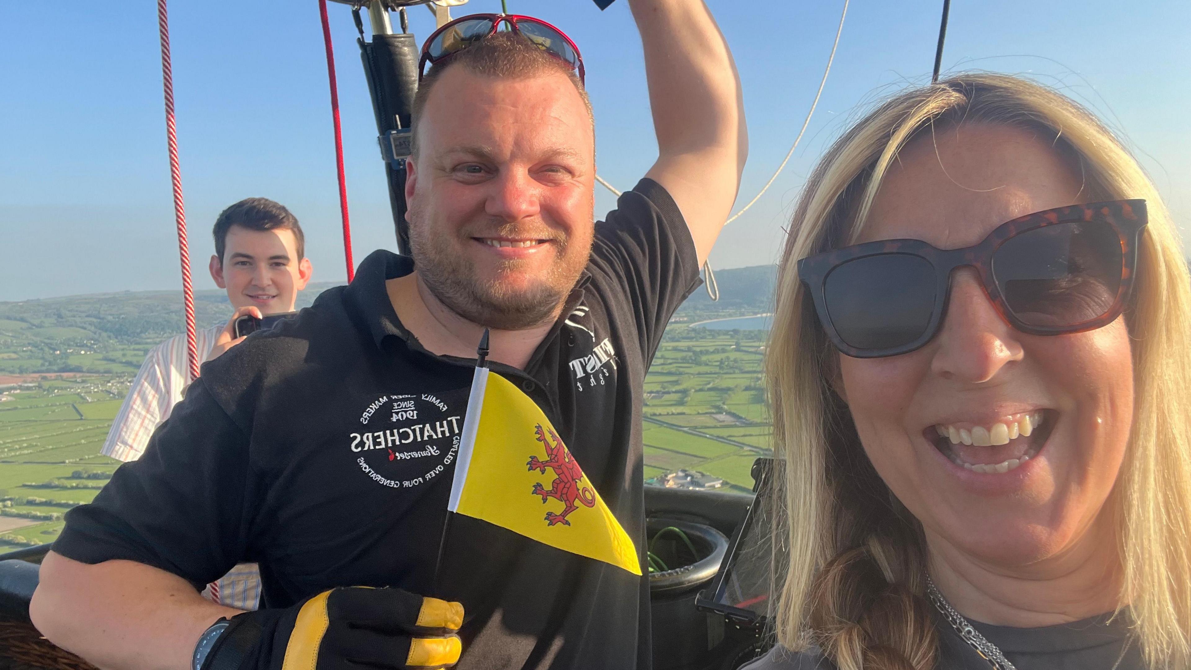 Three people are on a hot air balloon, they are all smiling at the camera. A man in the middle is holding up a small Somerset flag.