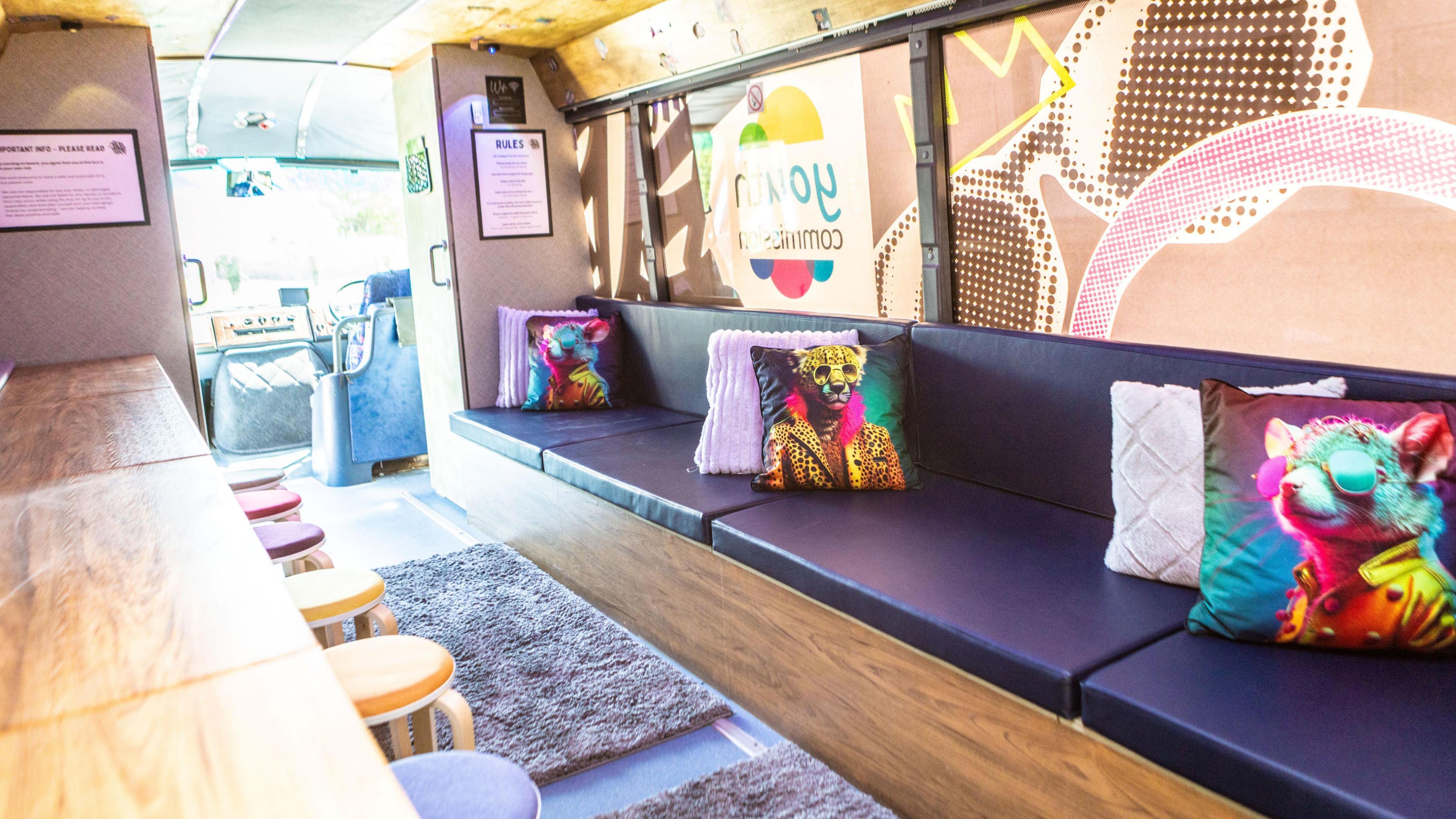 The image show the interior of the bus. It has a large seating area with a long bench with dark cushions and decorative pillows. There is also a desk area with lots of little stools underneath.