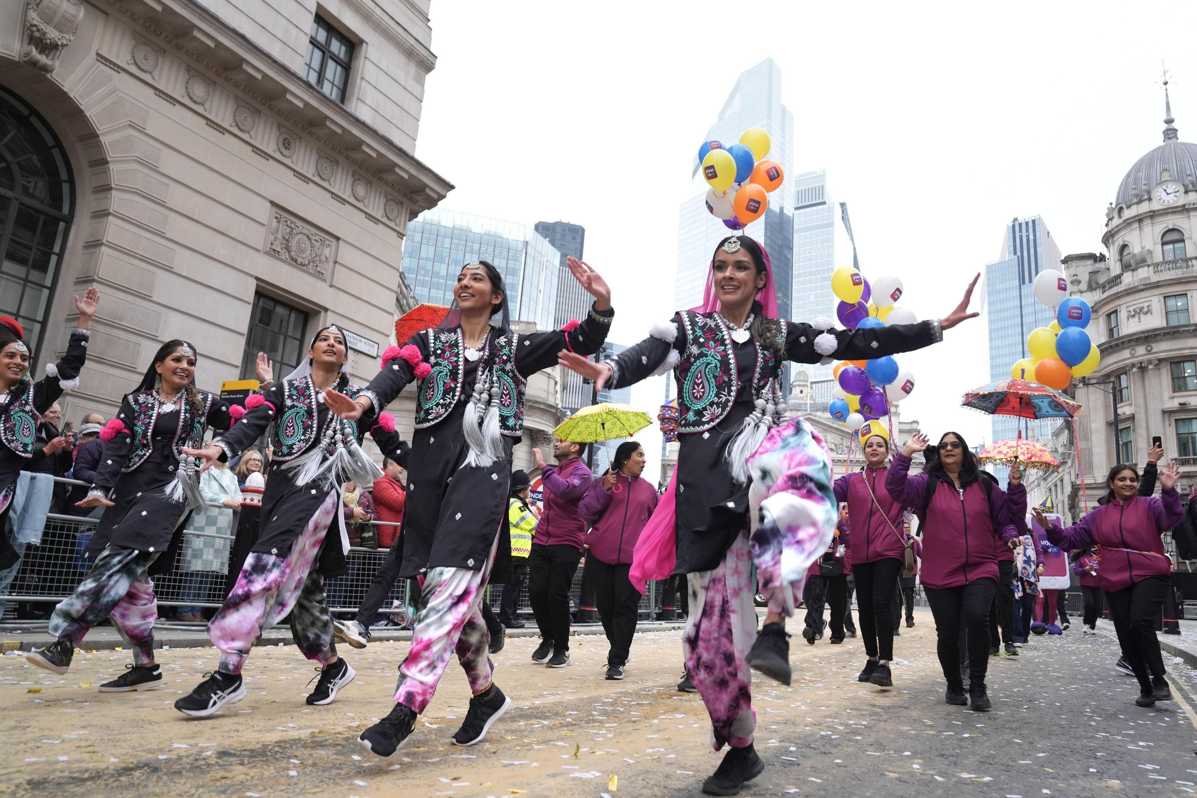 A vibrant group of women in colourful outfits performs a dance during the Lord Mayor's Show parade. They wear black vests with intricate embroidery and patterned trousers. Behind them, people carry umbrellas and balloons.