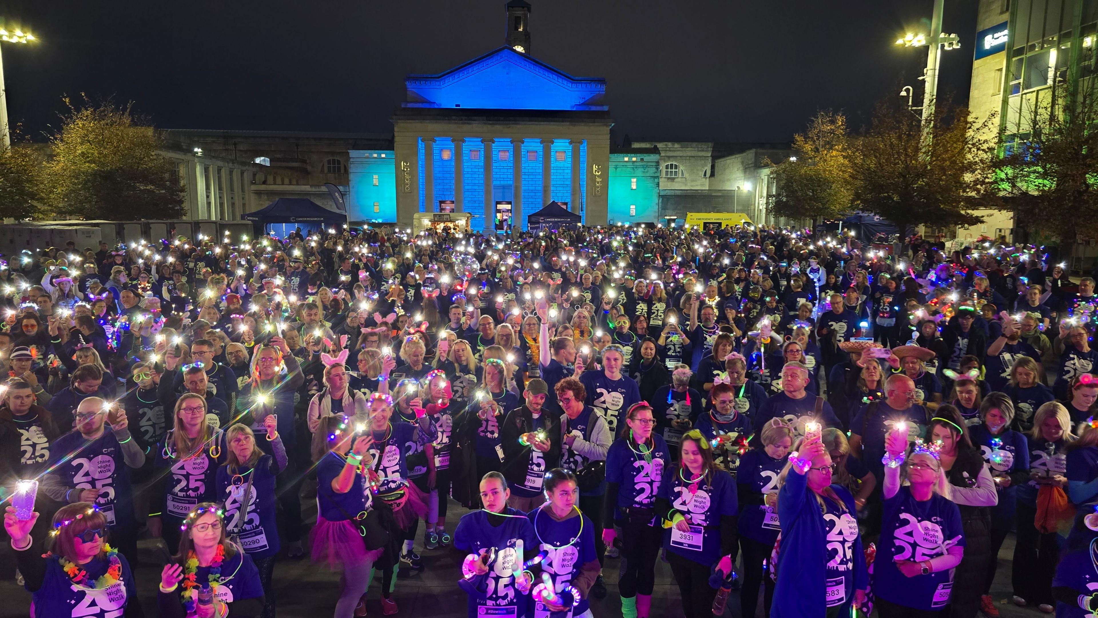 Hundreds of people holding up mobile phone torches and wearing matching event t-shirts with 2025 on them. Most of the people are wearing fairy lights and glow-in-the-dark necklaces. The classical columns of Southampton Guildhall are illuminated blue in the background.