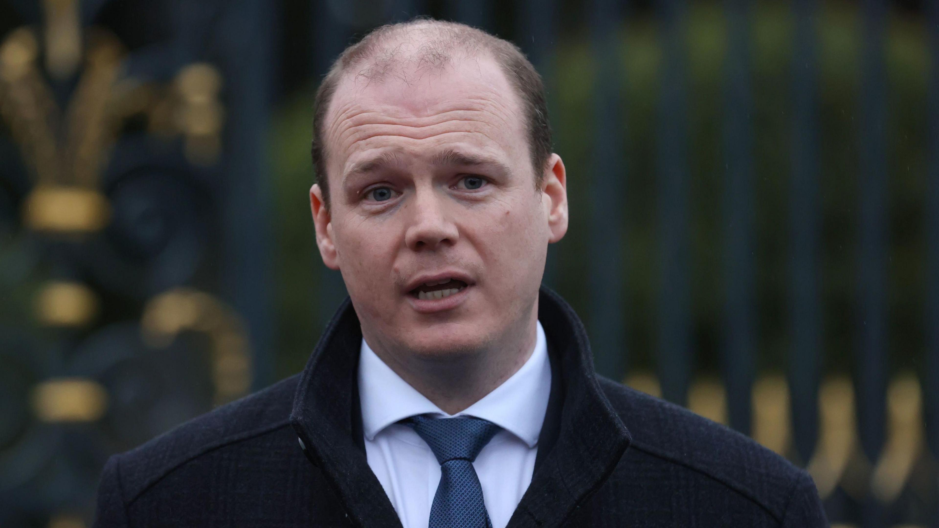 Gordon Lyons stands in front of a black fence, he wears a white shirt and blue tie with a black coat.