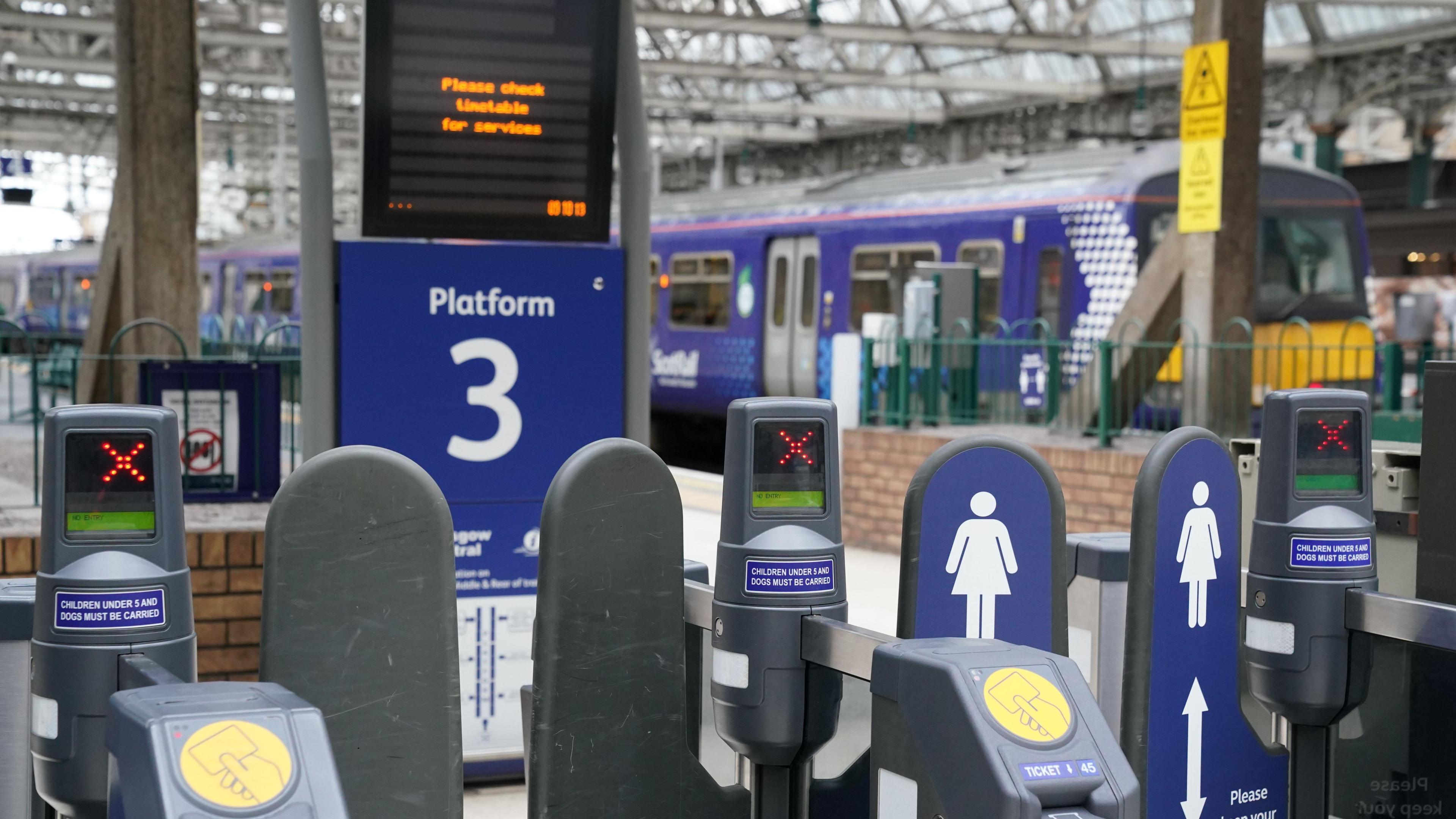 Ticket barriers at a train station, with a train in the background, and electronic departure boards displaying information