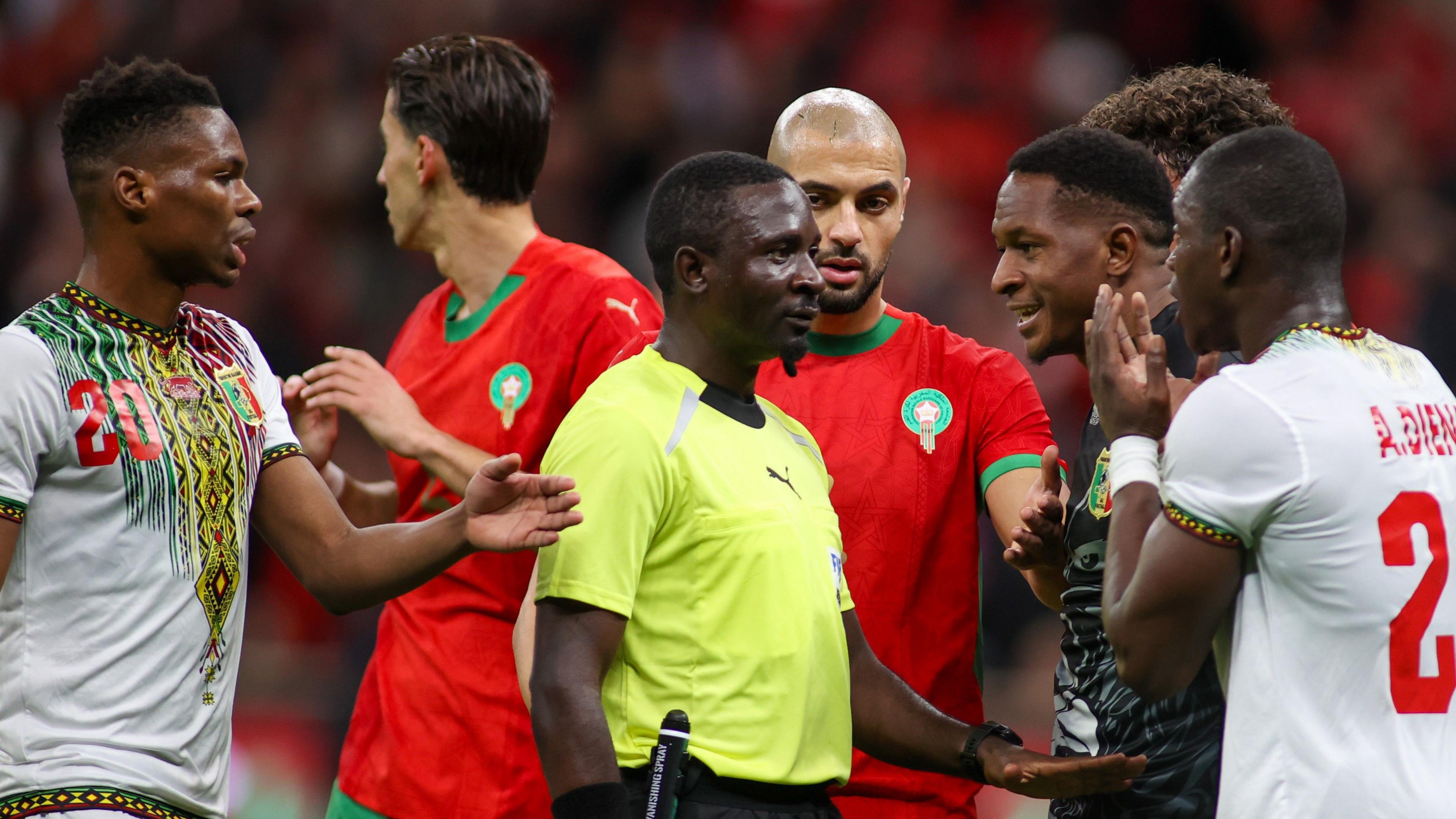 Cameroonian ref Abdou Abdel Mefire seen from waist up surrounded by a group of six protesting players during the match between Morocco and Mali at Afcon 2025. Mefire, who is looking from left to right in the image, is wearing a dayglo yellow short-sleeved top and is gesturing with his left hand with the hand held flat, palm down, to indicate that people should calm down. Two Mali players are right in front of him, including one with both hands up in front of his face to indicate disbelief
