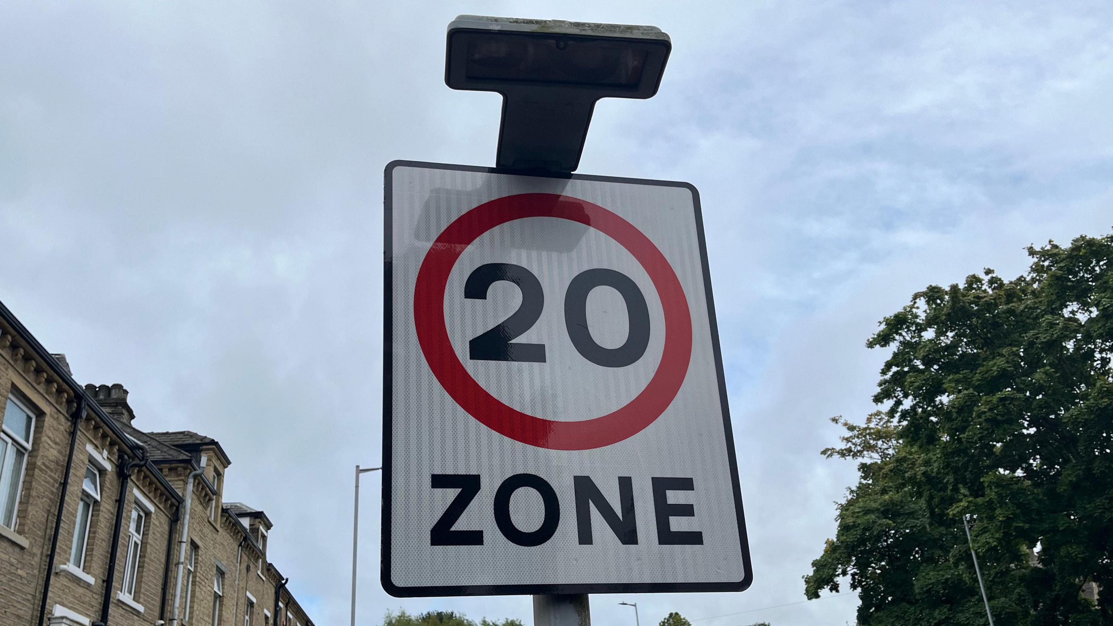 A white road sign that indicates a 20mph zone with a 20 written in black which has been encased in a red circle. Houses can be seen to the left of the sign.