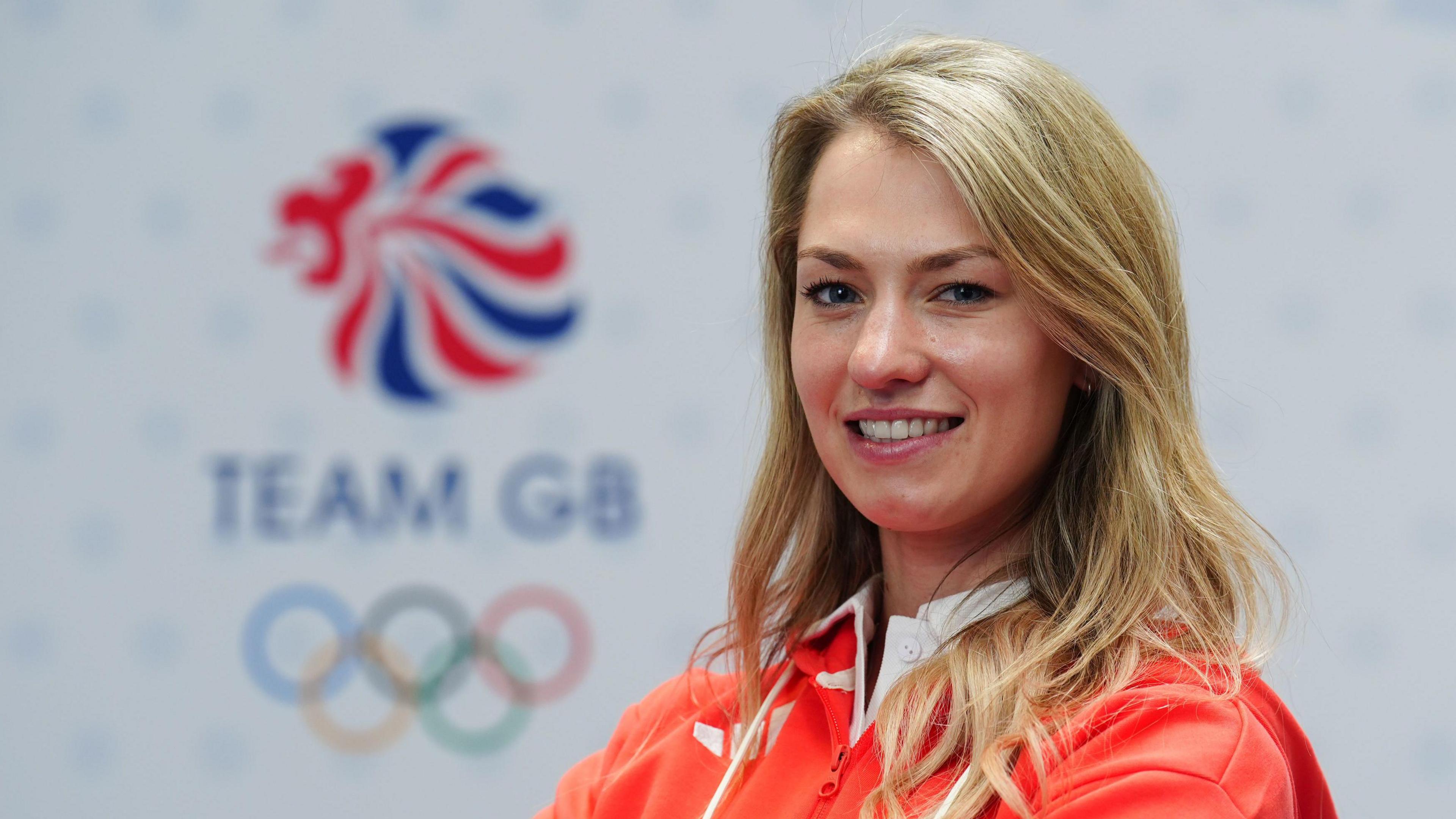 A smiling Lottie Fry wears the Team GB kit and poses for a photograph in front of the Team GB Olympics logo.