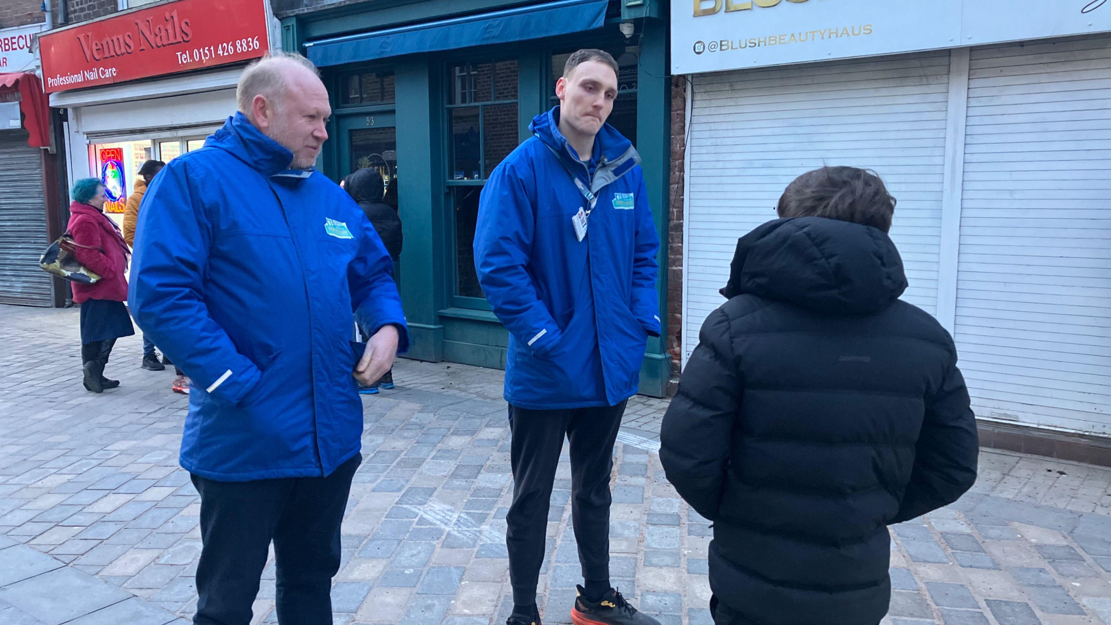 Three people are in the image - two in blue jackets are youth workers. They are facing the camera, talking to a young person in a black puffa coat who is facing away from us. They are standing in a street with a parade of shops behind them.