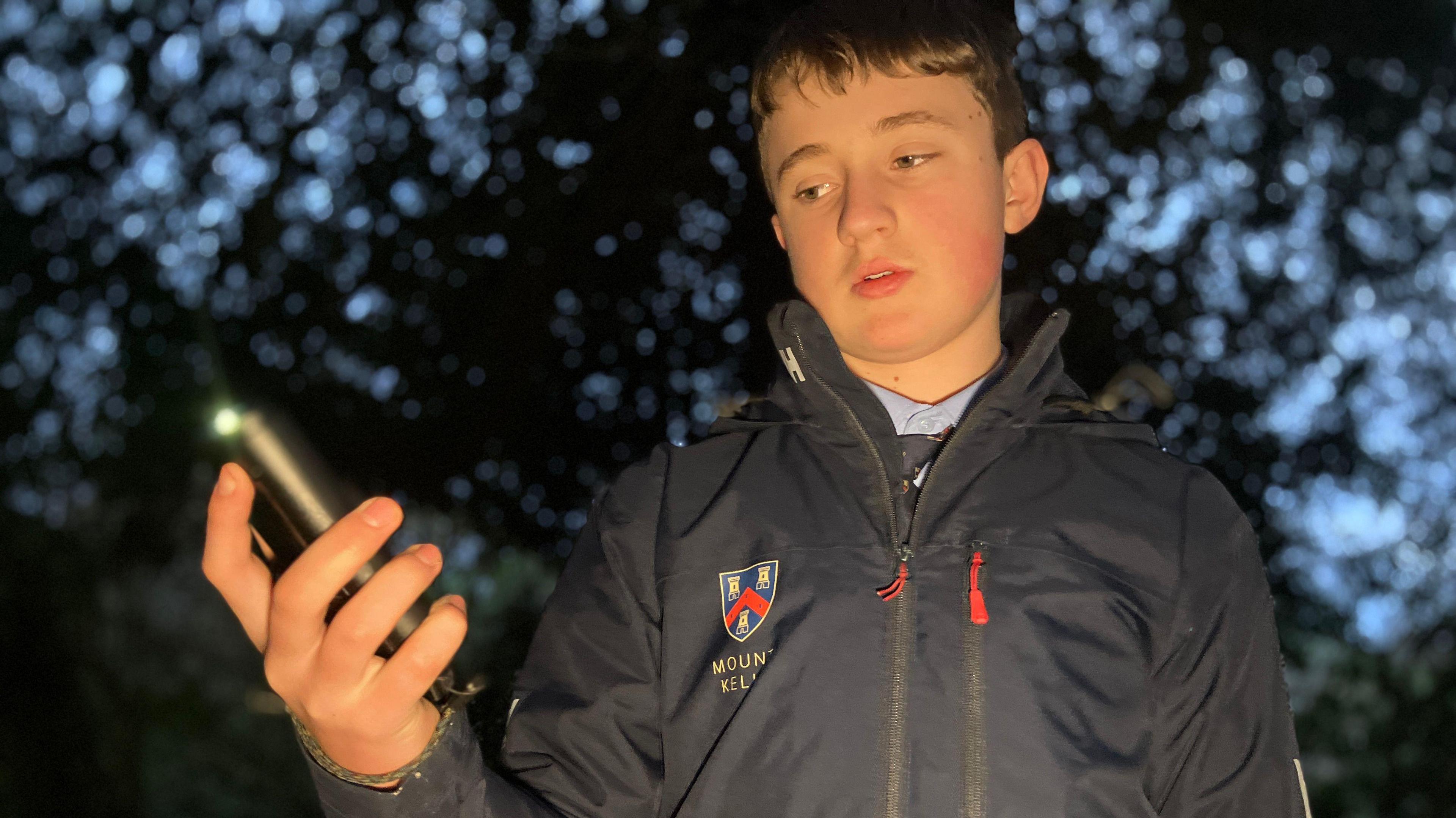 A boy wearing a black jacket has his hand in front of him holding a tracking device, he looks down toward the device.