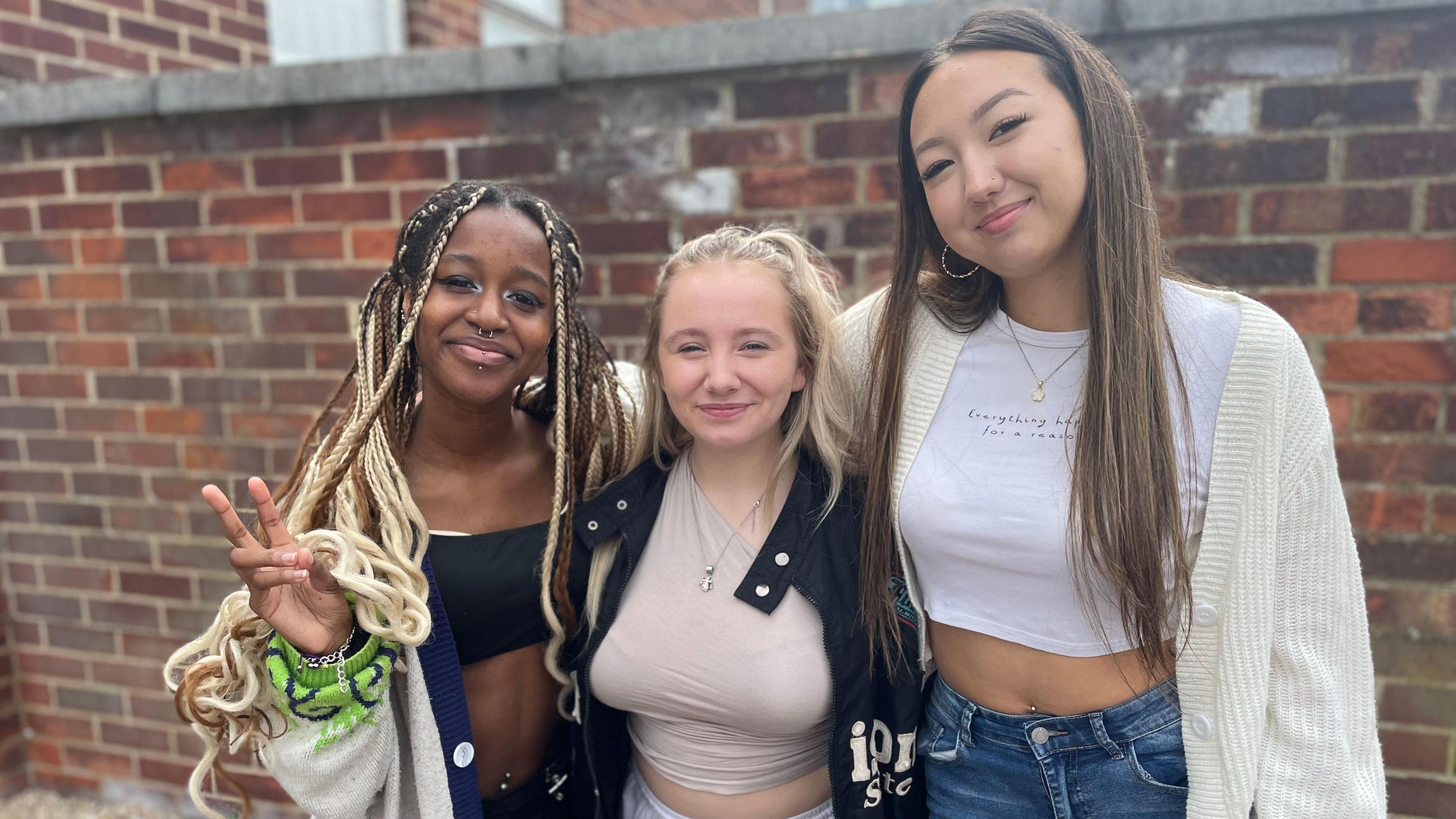 Three 18-year-old female students stand smiling at the camera. They are in front of a wall.