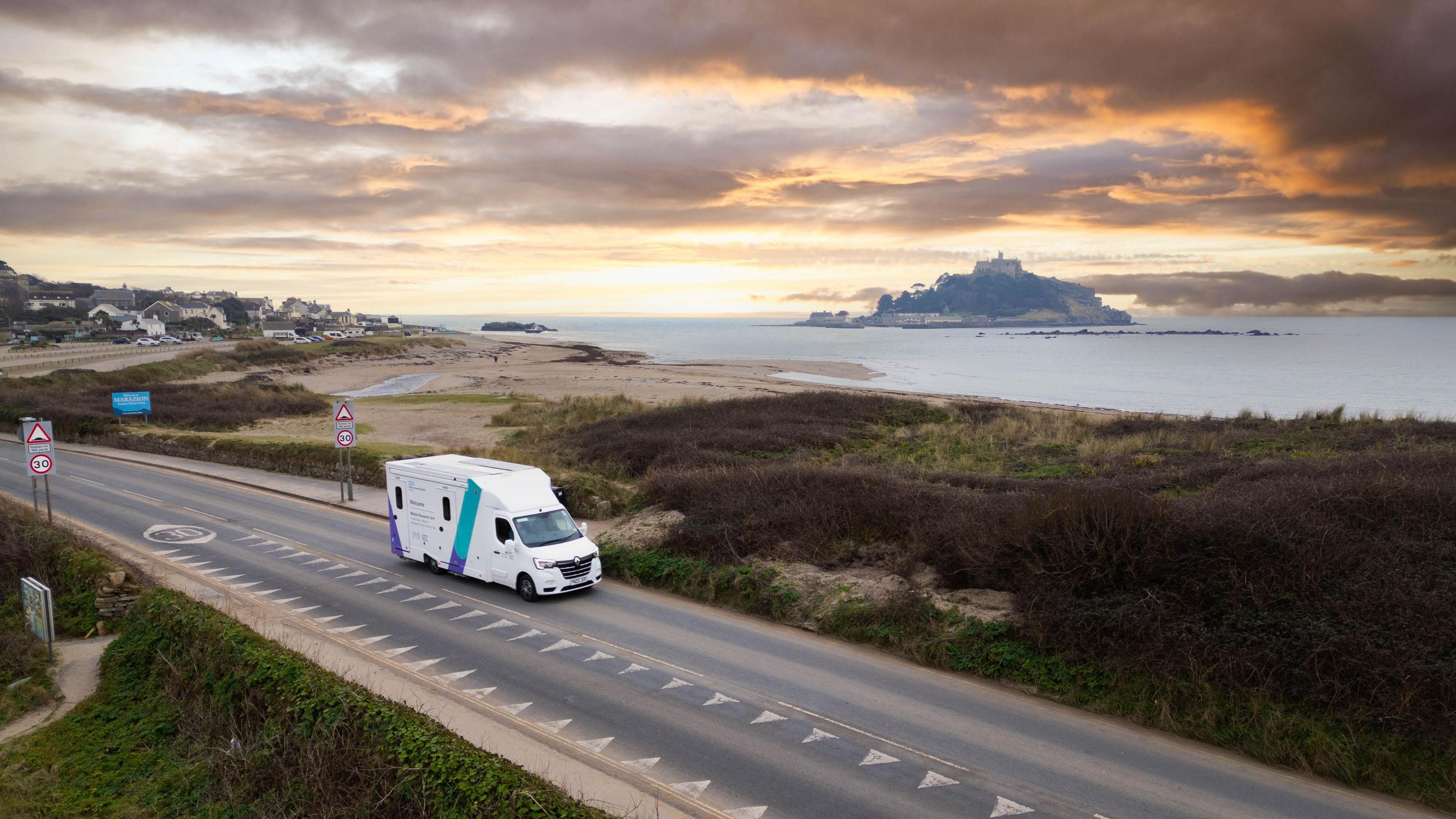 A drone shot showing the mobile research unit van driving along a road with the sea and St Michael's Mount in the background.