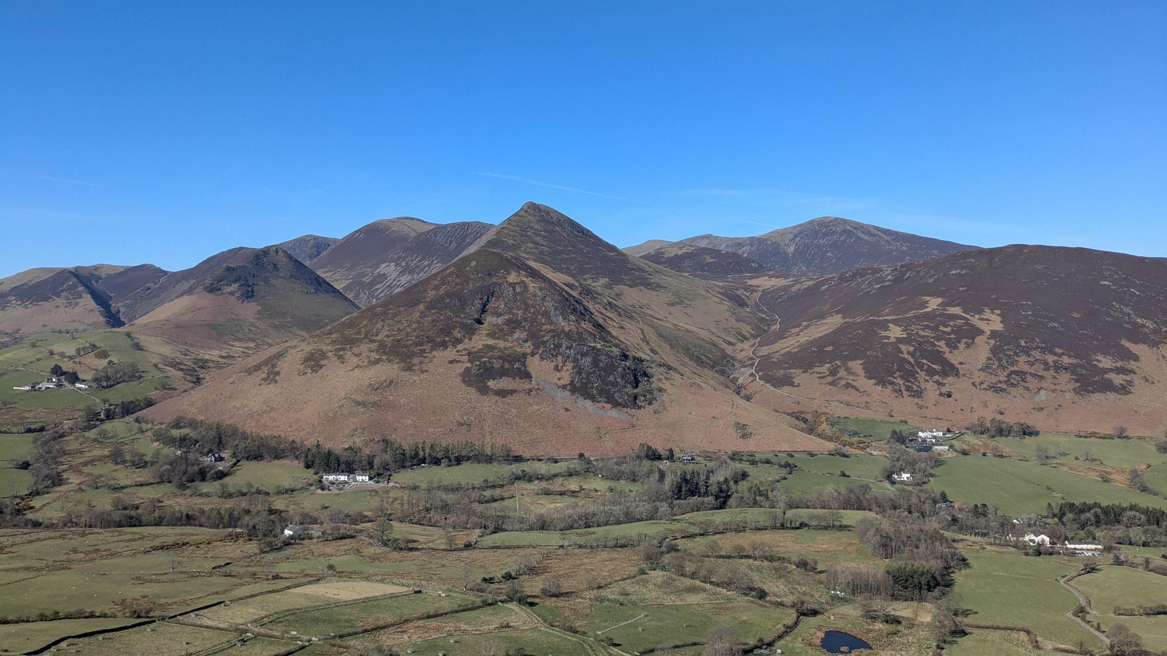 A general view of the fells taken from Cat Bells near Keswick in the Lake District. The green, undulating valley is sectioned by walls and hedges, with groupings of trees dotted around. Behind it imposing brown mountains rise up into a series of triangular peaks beneath a clear, deep blue sky.