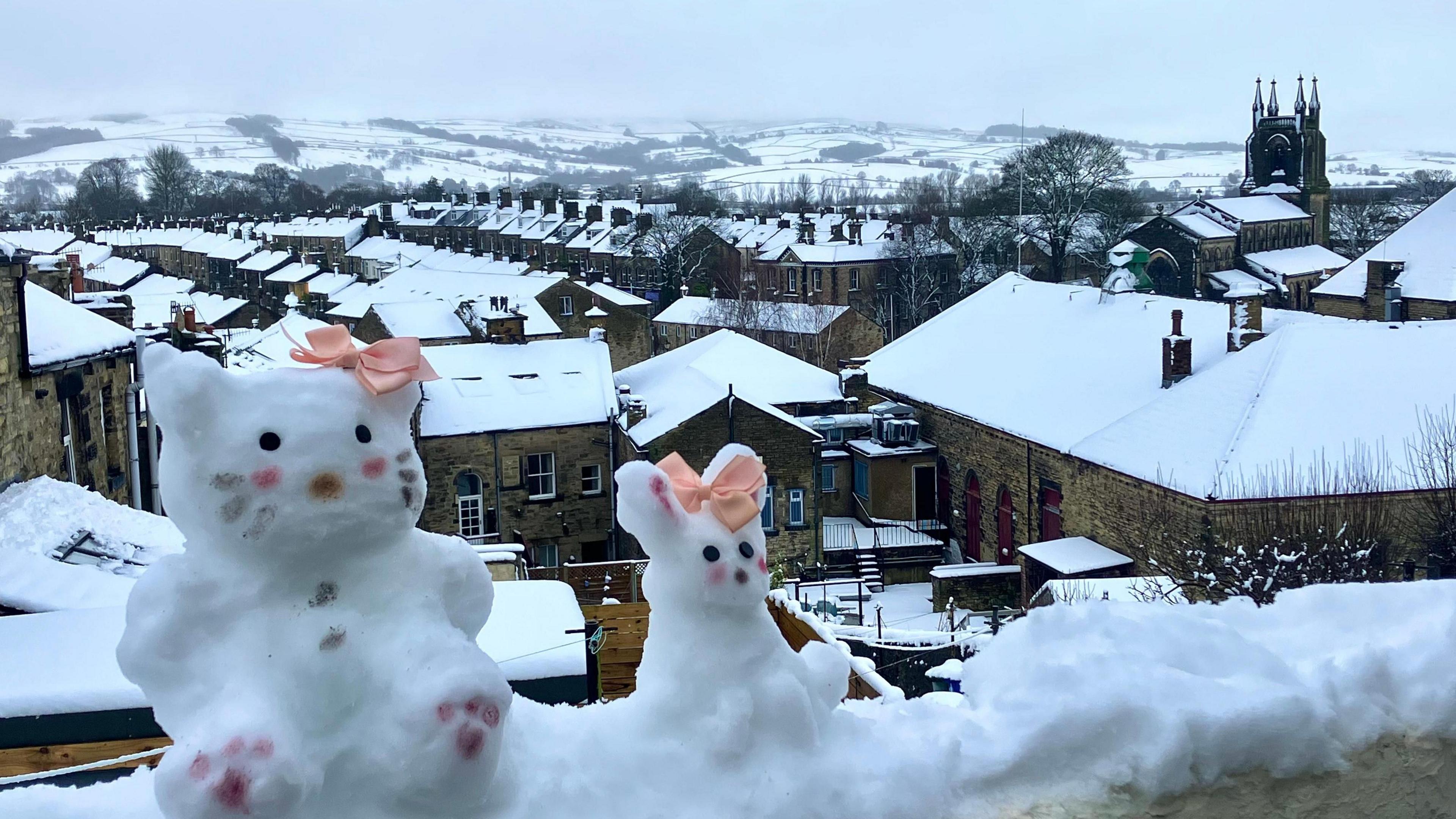 Two snow figures of bunnies, with eyes, nose, and pink dots on their cheeks, and pink ribbons on their heads, perch atop a wall overlooking snow-covered roofs of Skipton, North Yorkshire