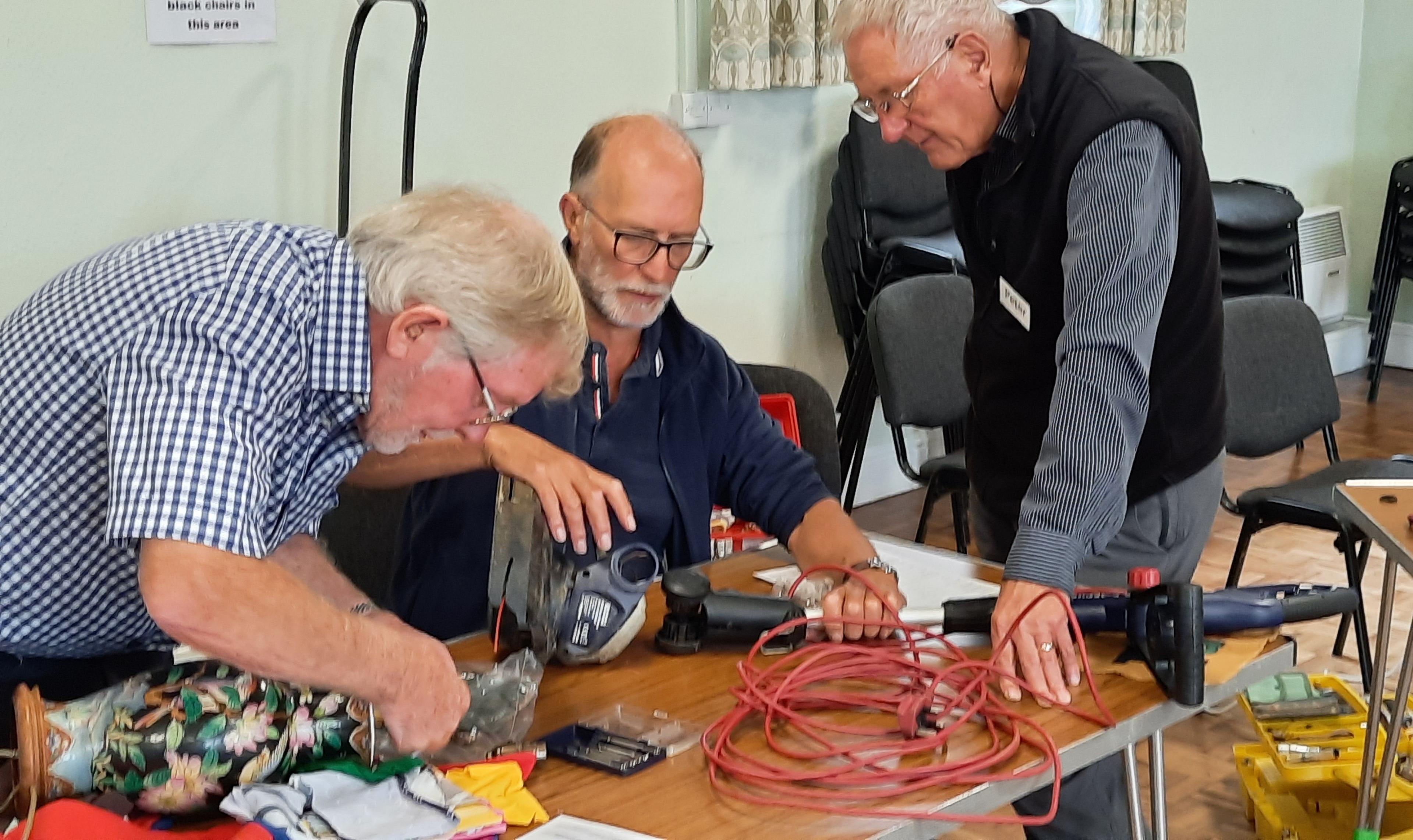 Three men with grey hair gather around a table with different tools on it. The man on the left holds a black vase with floral patterns on it while the other two men inspect a vacuum cleaner.