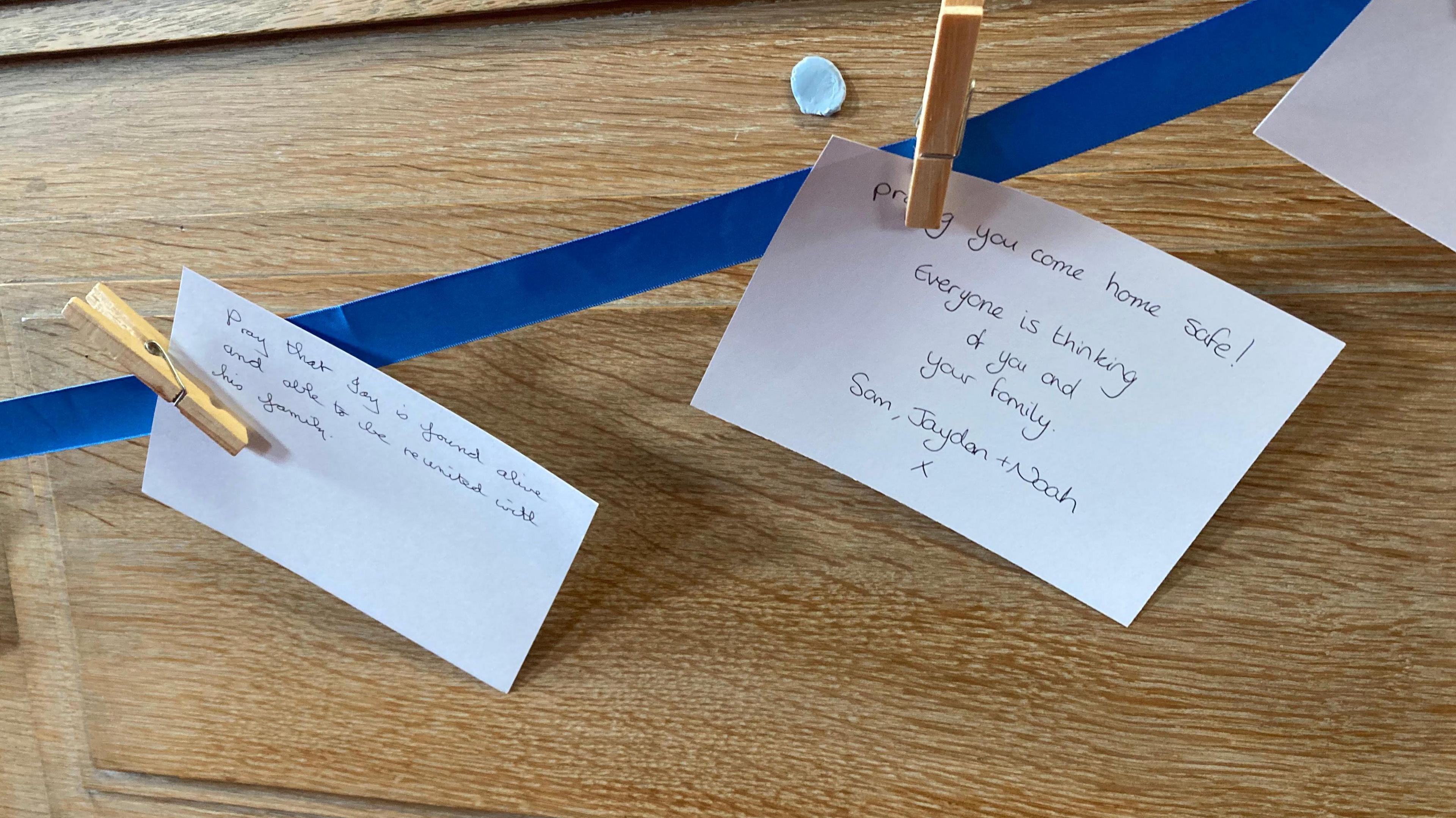 Two pieces of paper are clipped to a blue ribbon with a peg. One reads pray that Jay is found alive and another reads pray you come home safe. Everyone is thinking of you and your family.