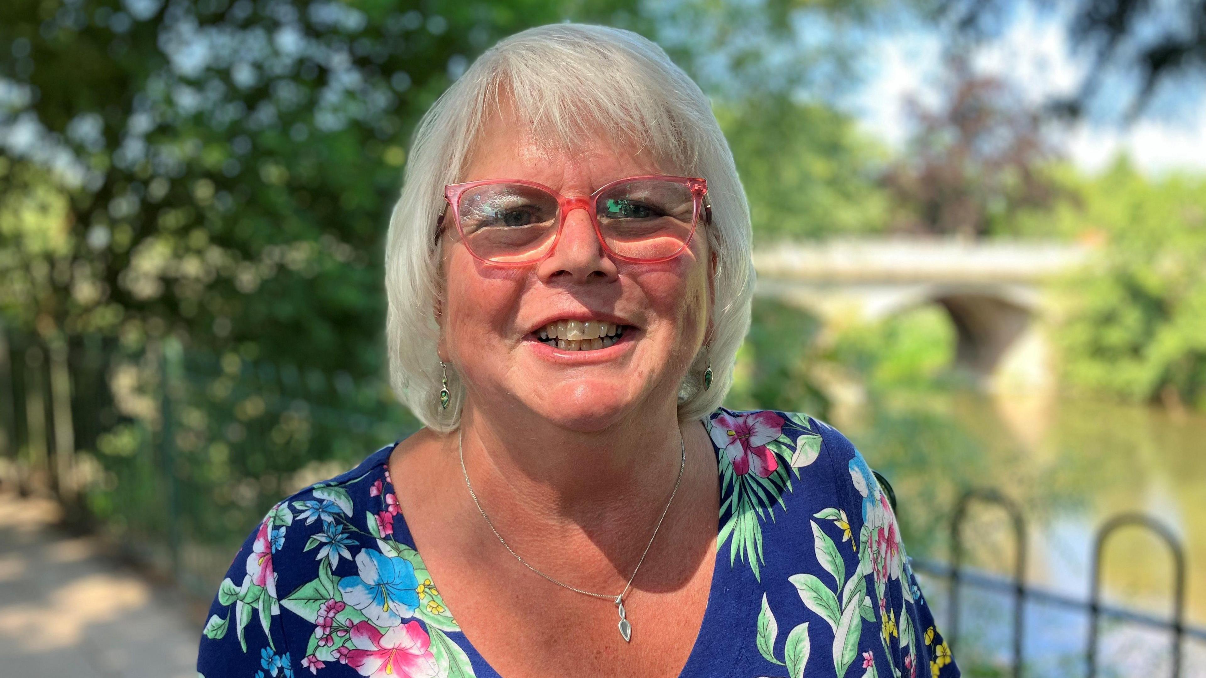 A woman smiling at the camera. She is wearing red-rimmed glasses and a blue floral top, and standing by railings along the river. 