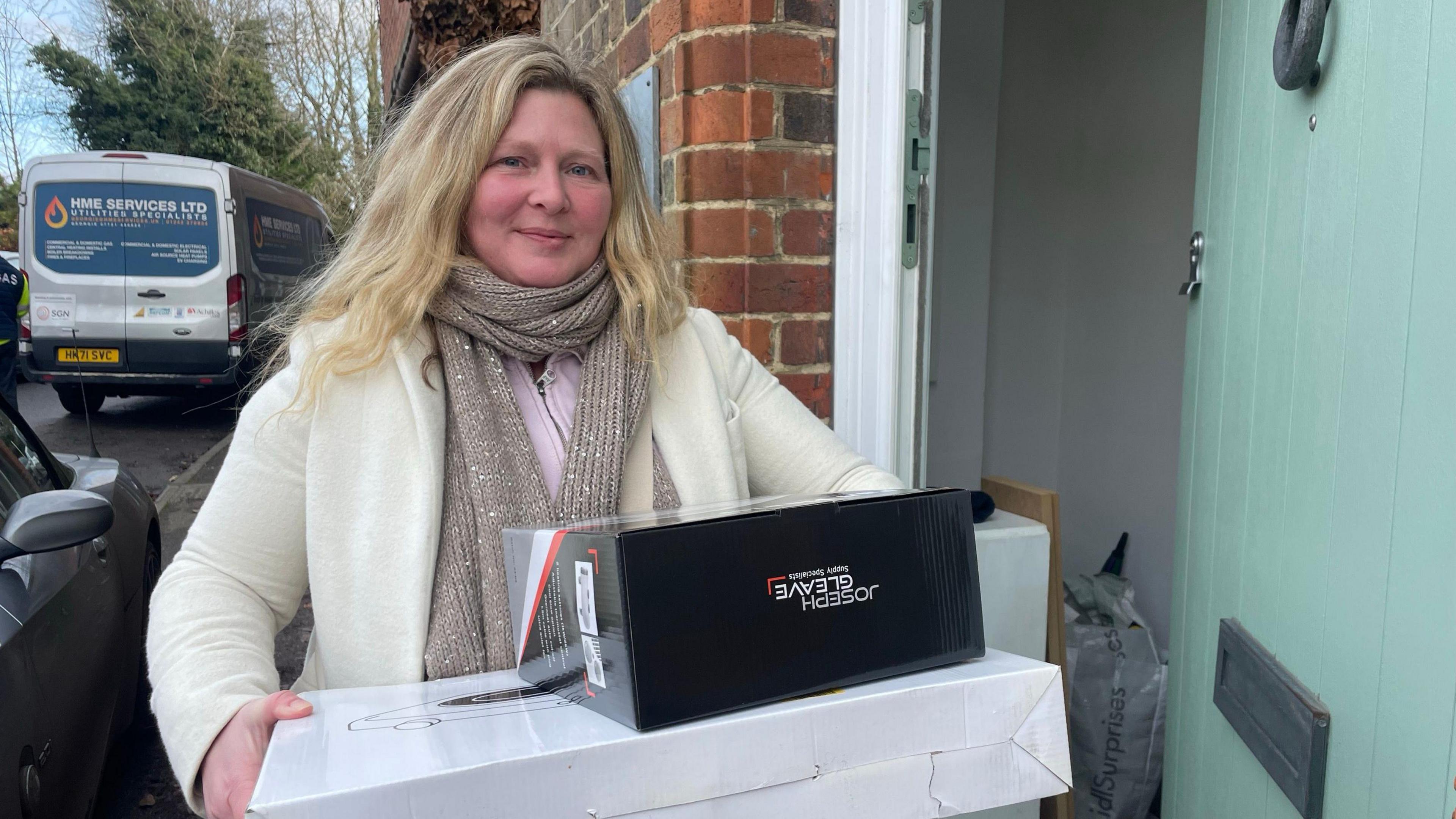 Terri Dempsey - a woman with blonde hair, wearing a white coat and grey scarf, holds a white box and a black box