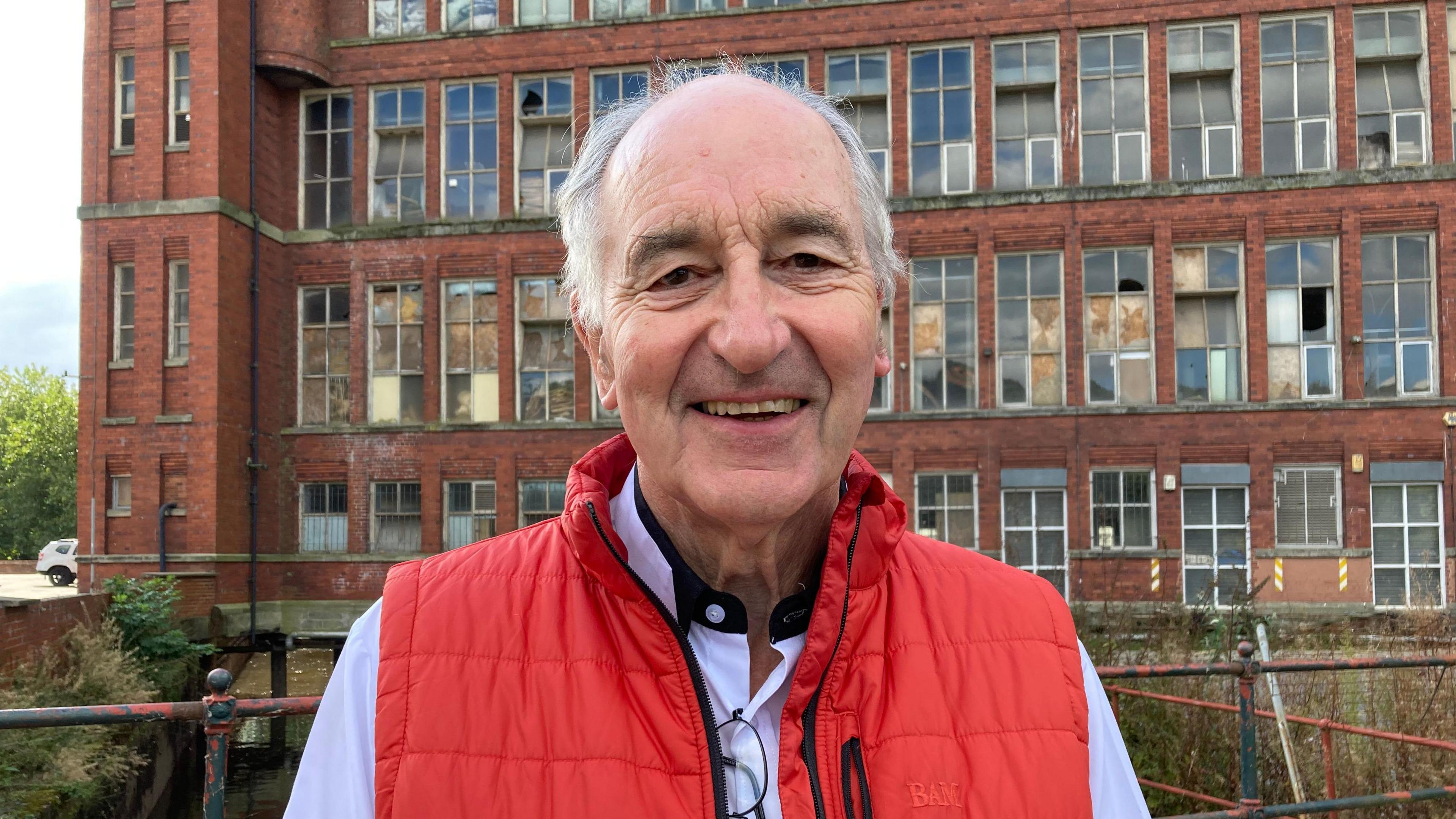 The picture is Derek Latham who is the chair of the Derbyshire Historic Buildings Trust. He is smiling and wearing a red, sleeveless coat and light blue shirt. Behind him are the old Belper Mills buildings