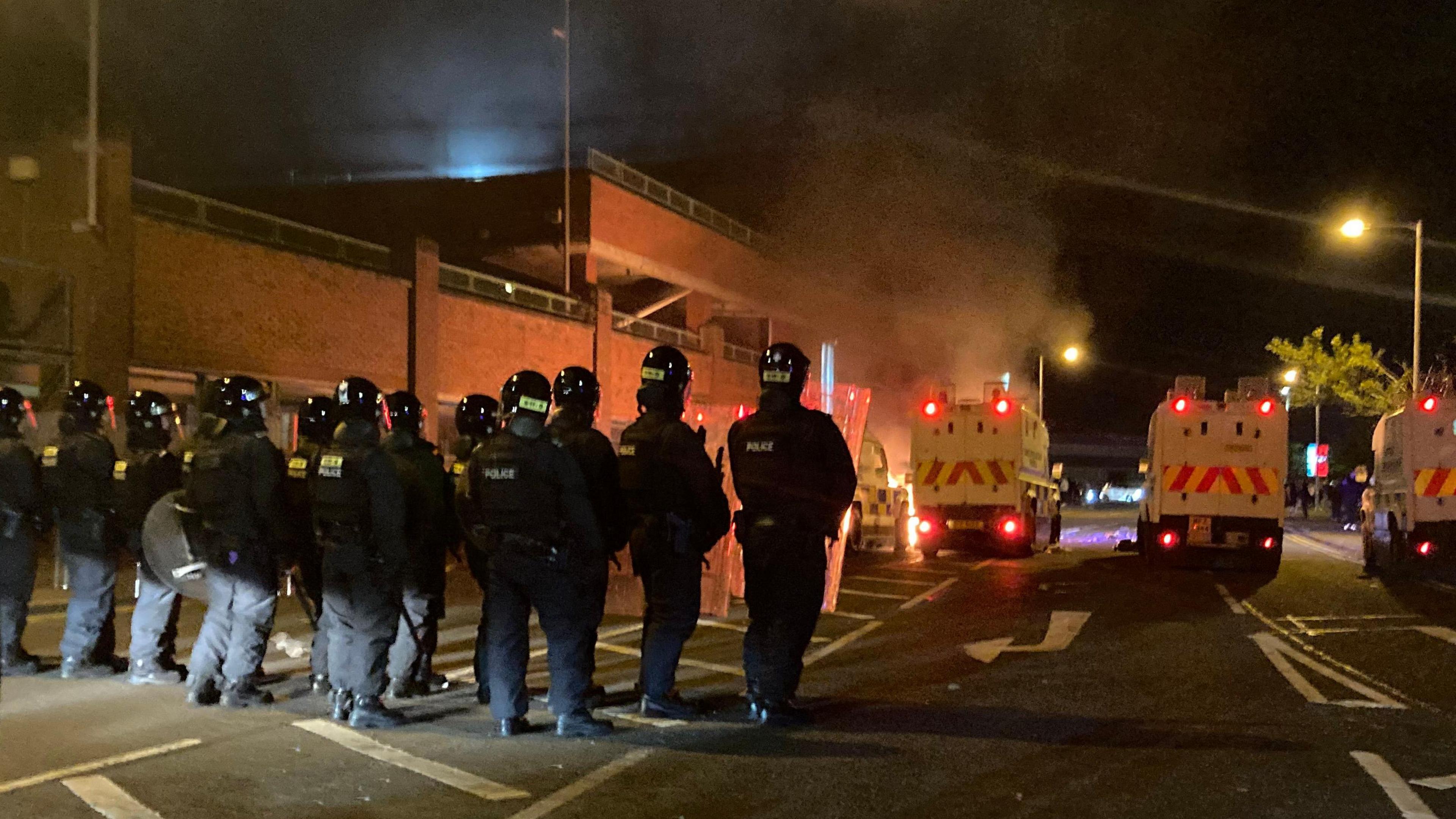 Row of police officers with their backs to camera, facing the police vehicles in their riot gear. Fire is in the distance. Police have shields.