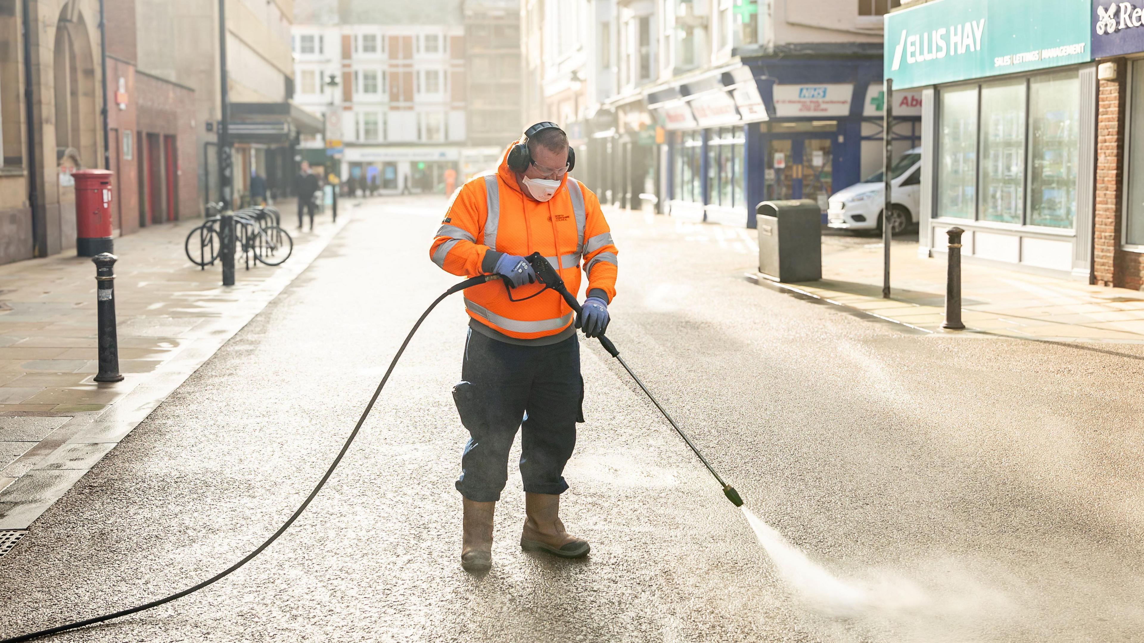 A man in an orange vest with a mouthguard and ear protectors on is jet washing a street.