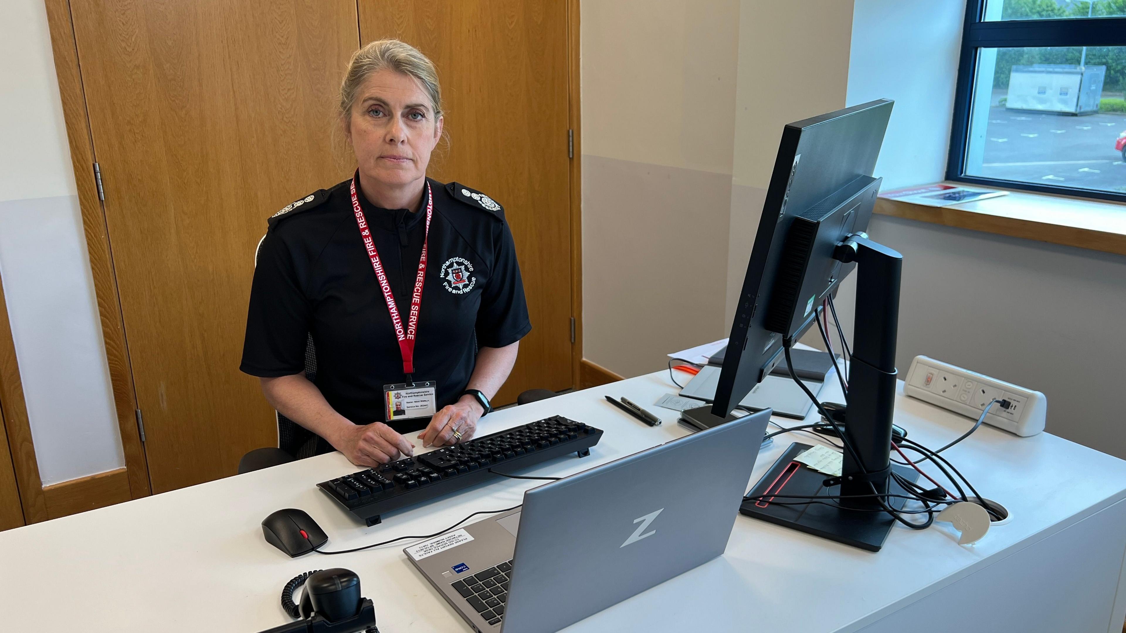 Northamptonshire Fire and Rescue Service chief fire officer Nikki Watson sitting behind a desk. She is wearing a black blouse and has an identity card on a red lanyard around her neck. There is a laptop, monitor and keyboard on the desk.