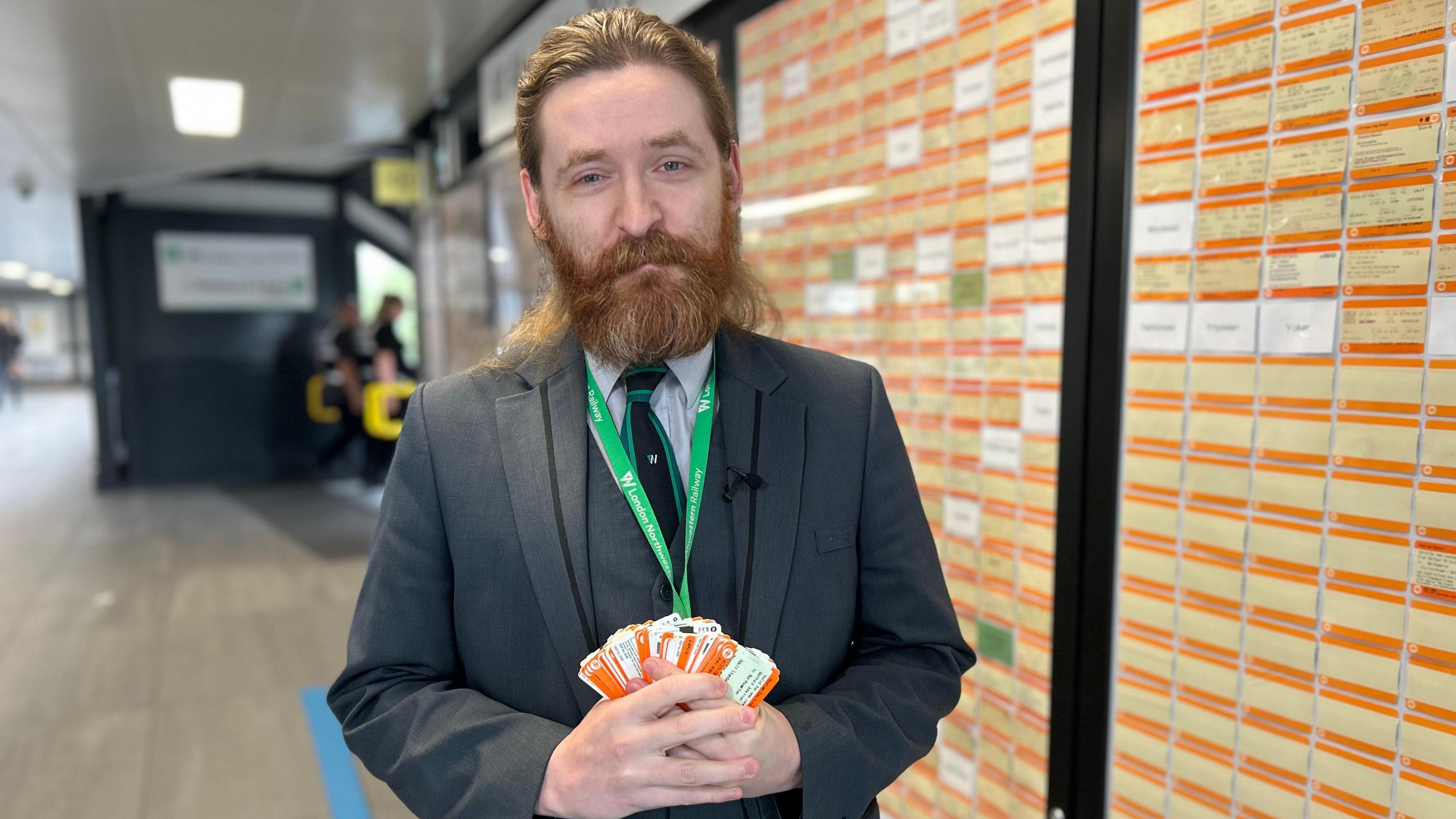 Elliott Badger with brown hair and a beard wearing a grey suit and a green lanyard holding a stack of orange rail tickets at Northampton railway station in front covered in orange train tickets