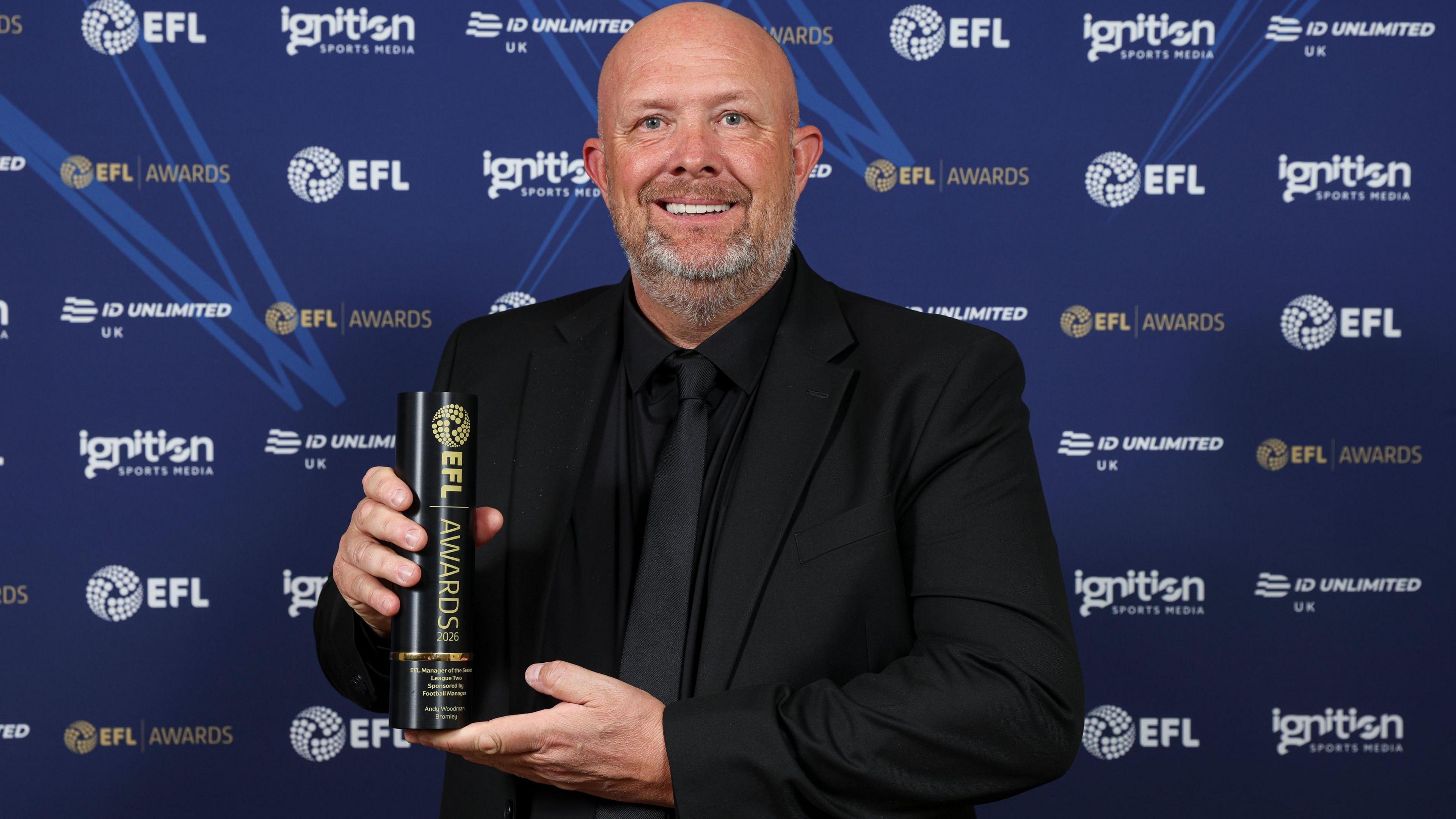 Andy Woodman poses with the League Two manager of the year trophy at the EFL Awards, dressed in an all-black suit, shirt and tie
