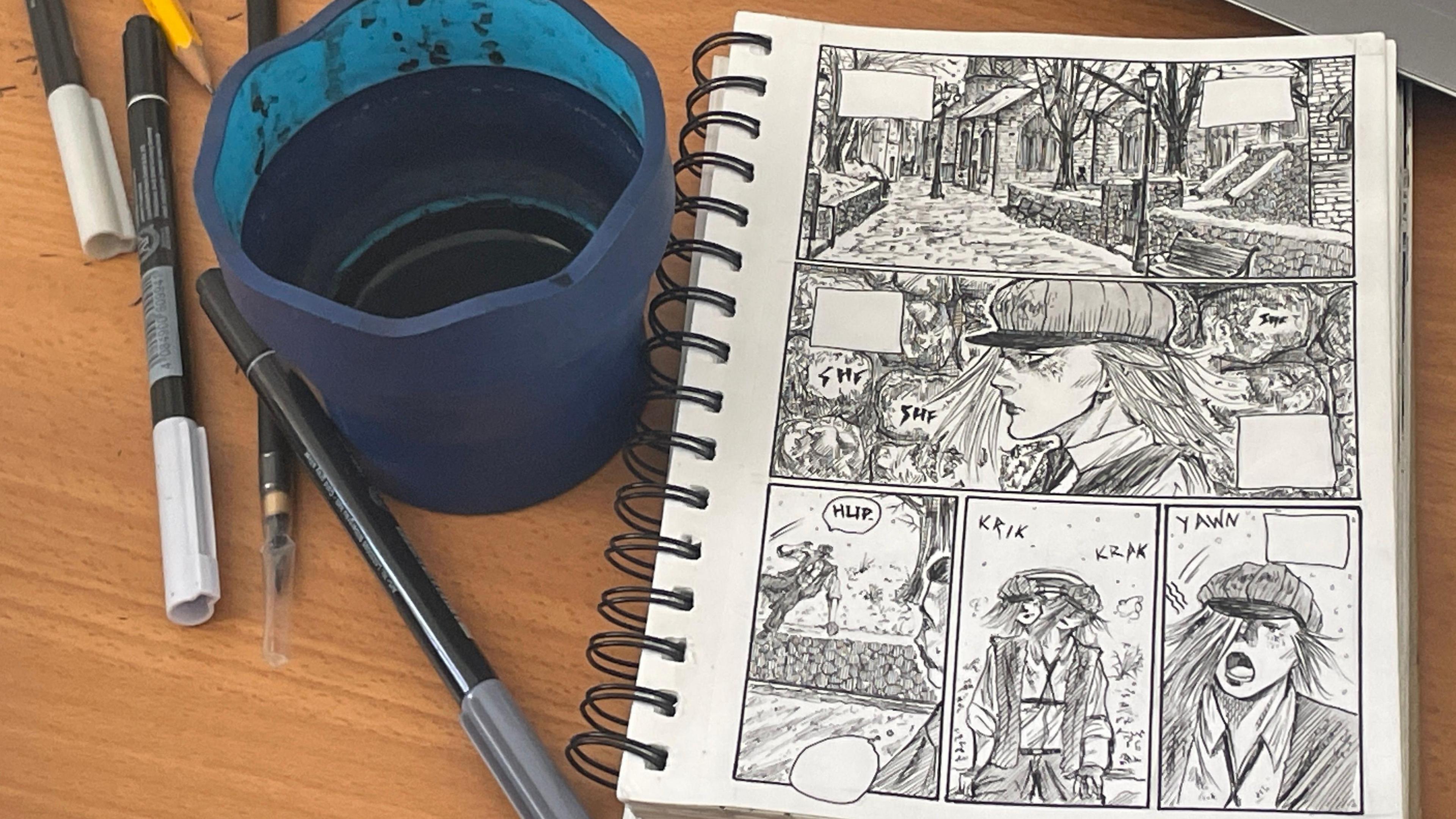 Aaban's notebook with sketches for his graphic novel sits on a desk, along with ink and pens and a cup that he washes paint brushes in.