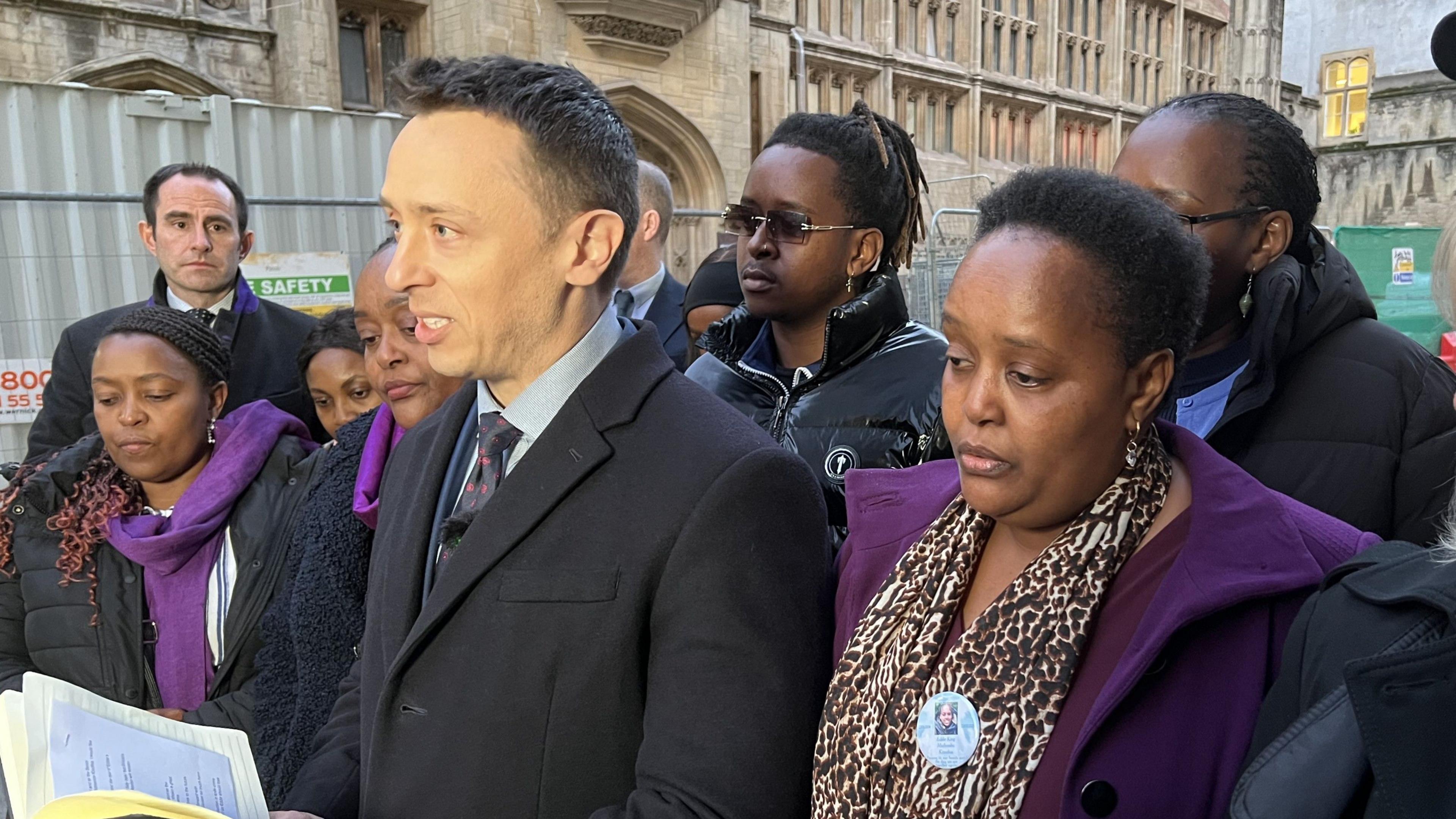 DCI Mark Almond delivering a statement to press outside court, reading from an open notebook in his hands. The family of Eddie Kinuthia, including his mum Irene, are surrounding him and look very solemn. They are all wearing a purple item of clothing, Eddie's favourite colour.