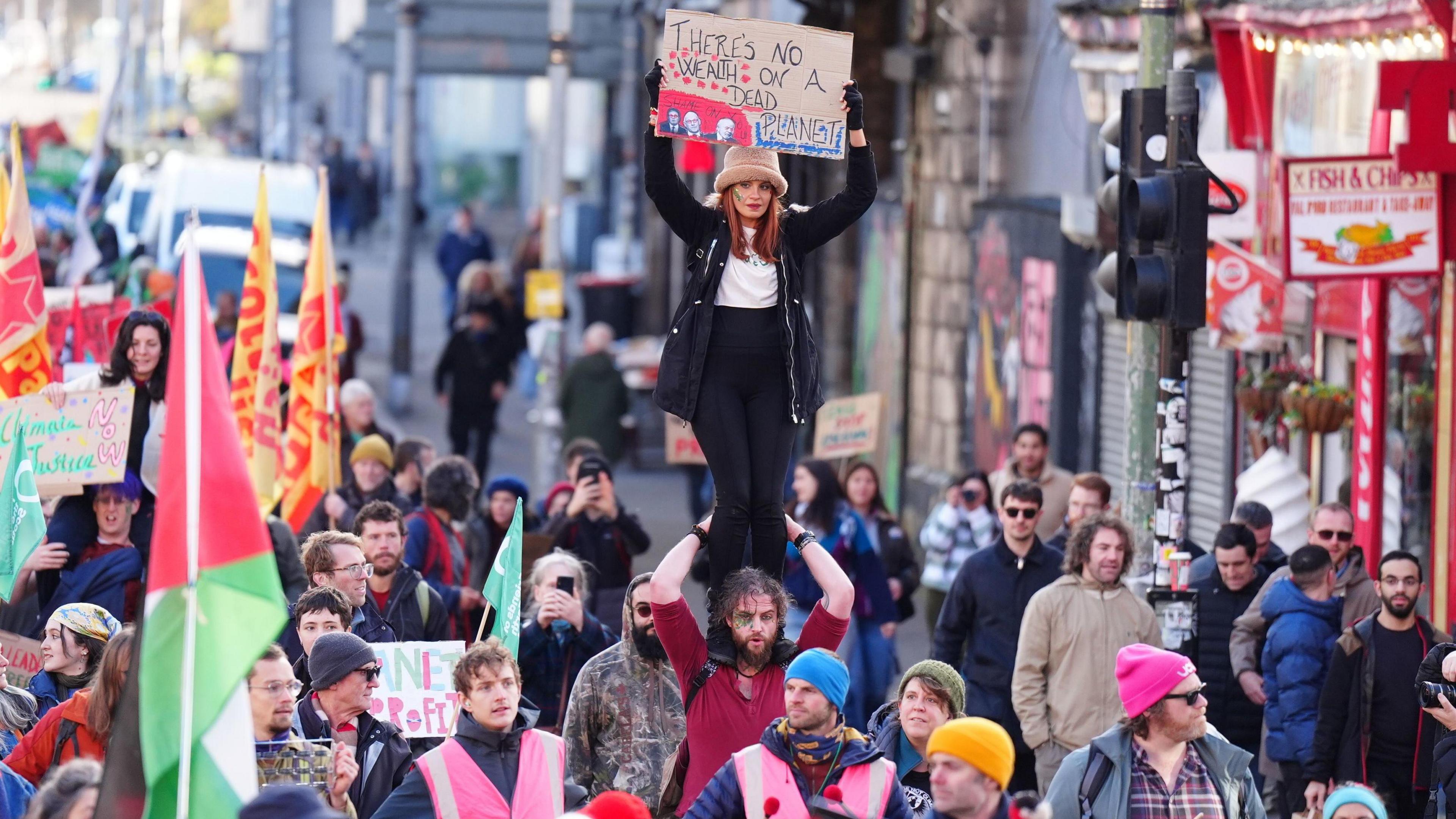 A large crowd of protesters marches down a city street. At the centre, a woman wearing a hat stands on someone’s shoulders and holds a sign above her head that reads, “There’s no wealth on a dead planet.” People around her carry flags and placards with various climate-justice messages. The street is busy with demonstrators, some in bright pink steward vests, and shops and buildings line the background.