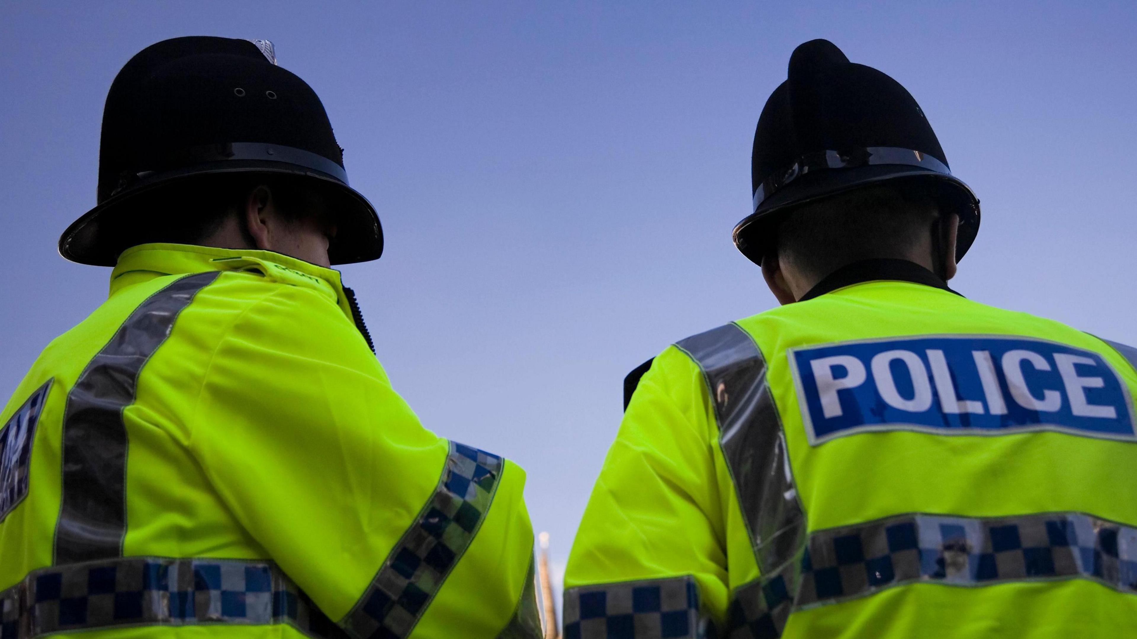 Back of two police officers wearing helmets and high visibility jackets with the word POLICE on the back. It is daylight