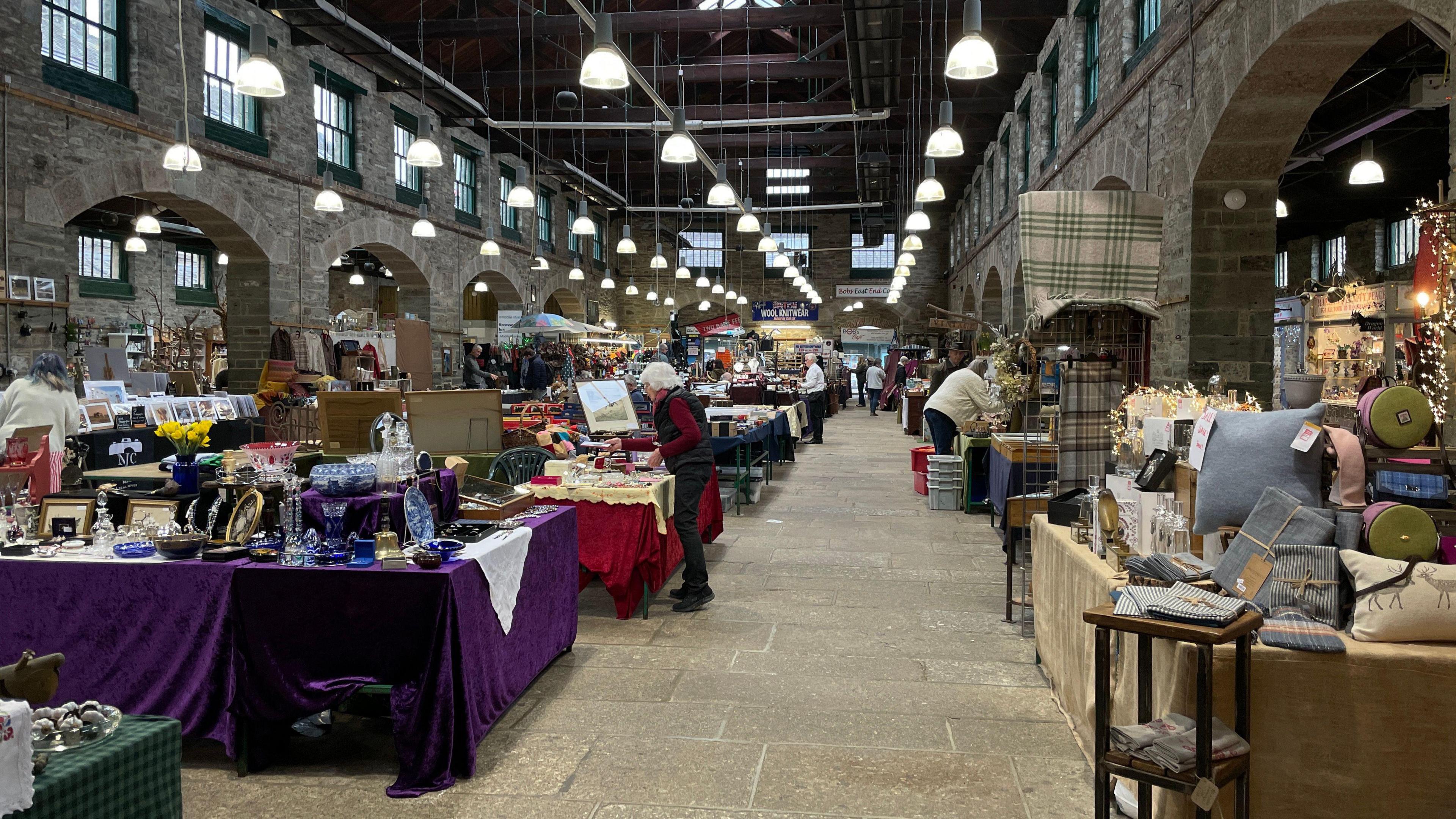 Inside Tiverton Pannier Market. A number of stalls are scattered around the market. Stallholders are making last minute adjustments to their displays. The stone hall is filled with goods including antique items cushions and household goods. Lights hang from the ceiling, illuminating the hall. 