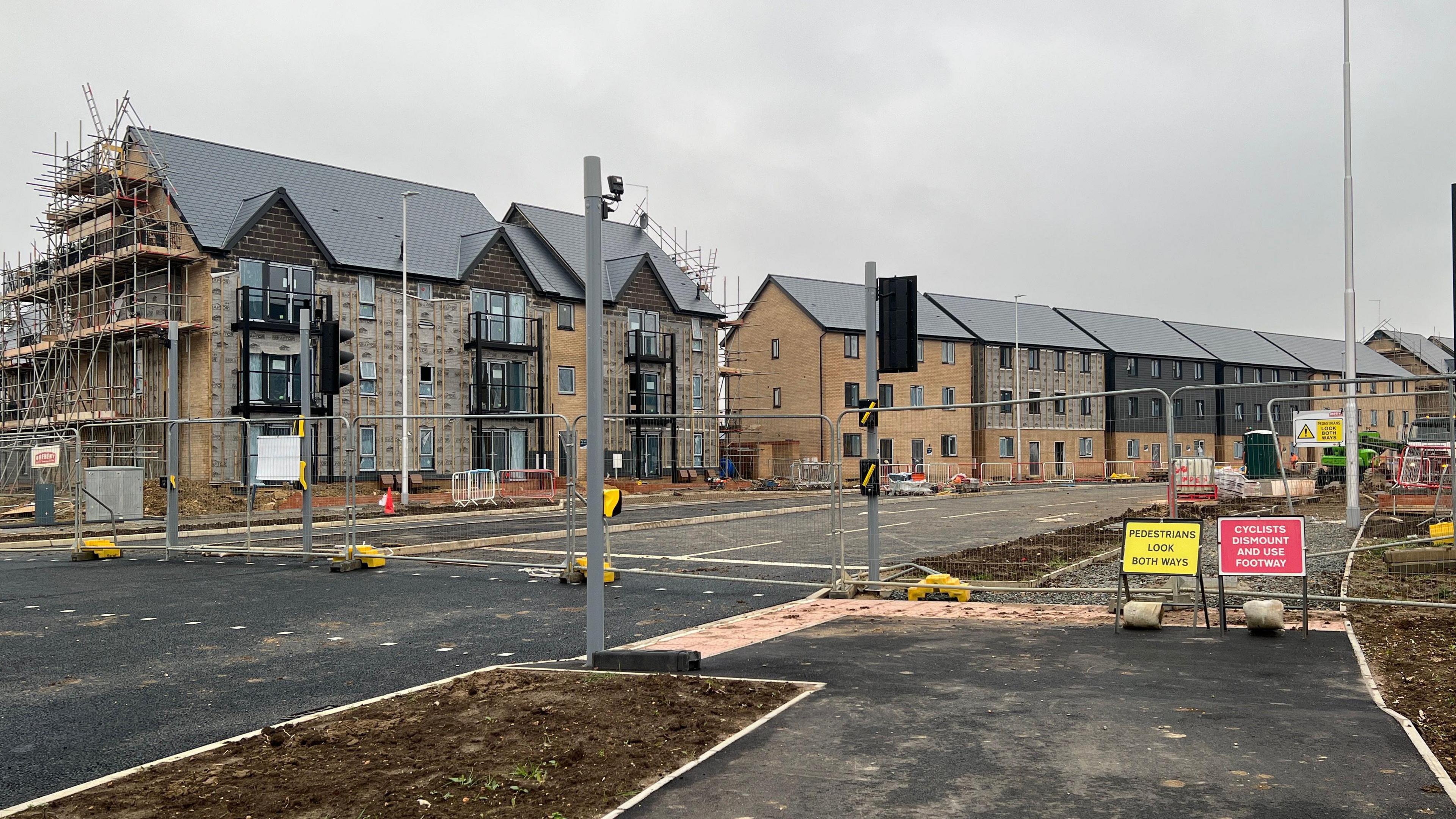 Under-construction row of houses. There is a metal security fence across an unfinished road. There are two small road signs - one is yellow and it is next to a red one. 