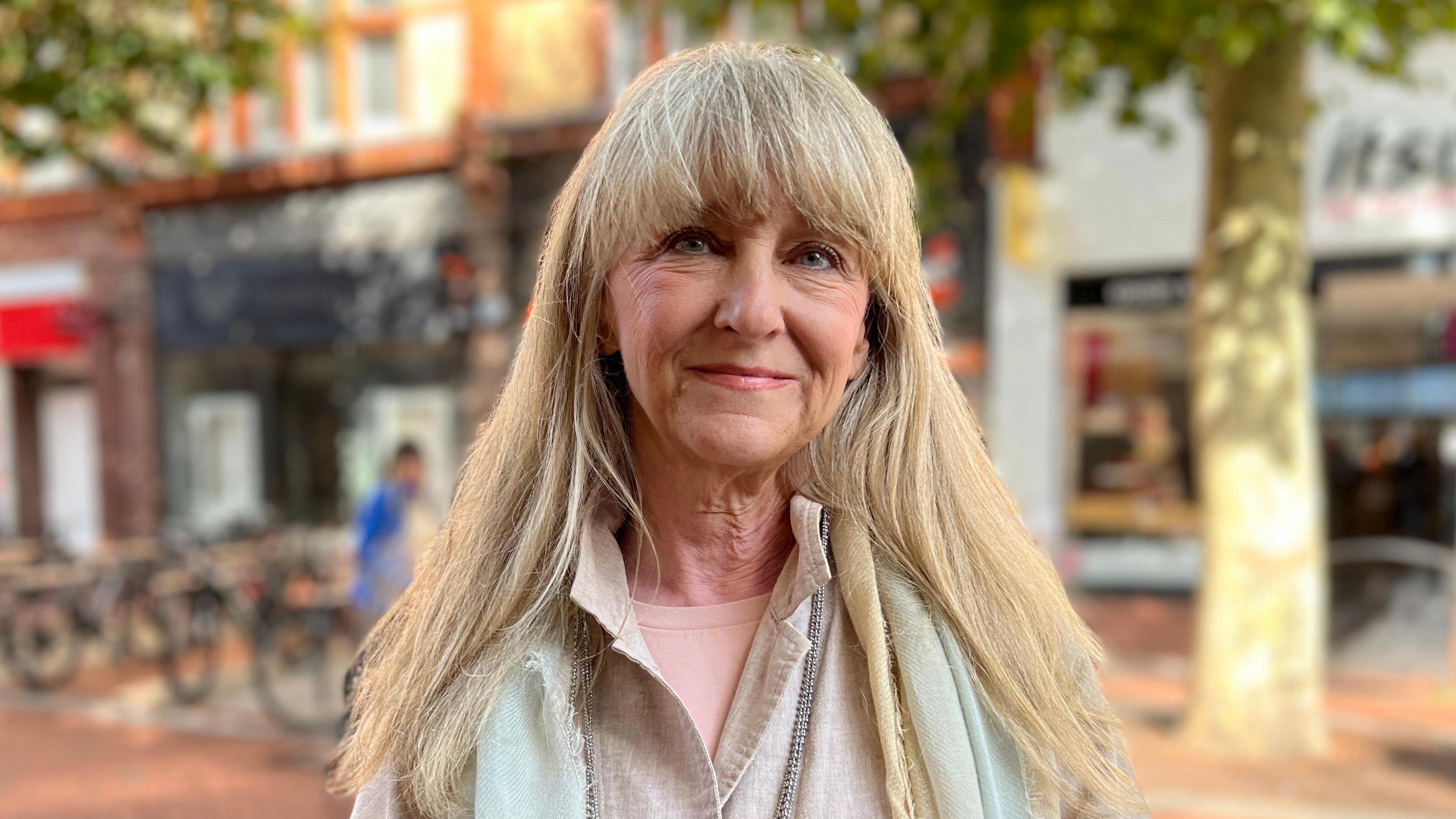 Karen Rowland stands facing the camera on Reading's Broad Street. She is wearing a cream coloured shirt with a white scarf and has long blonde hair.