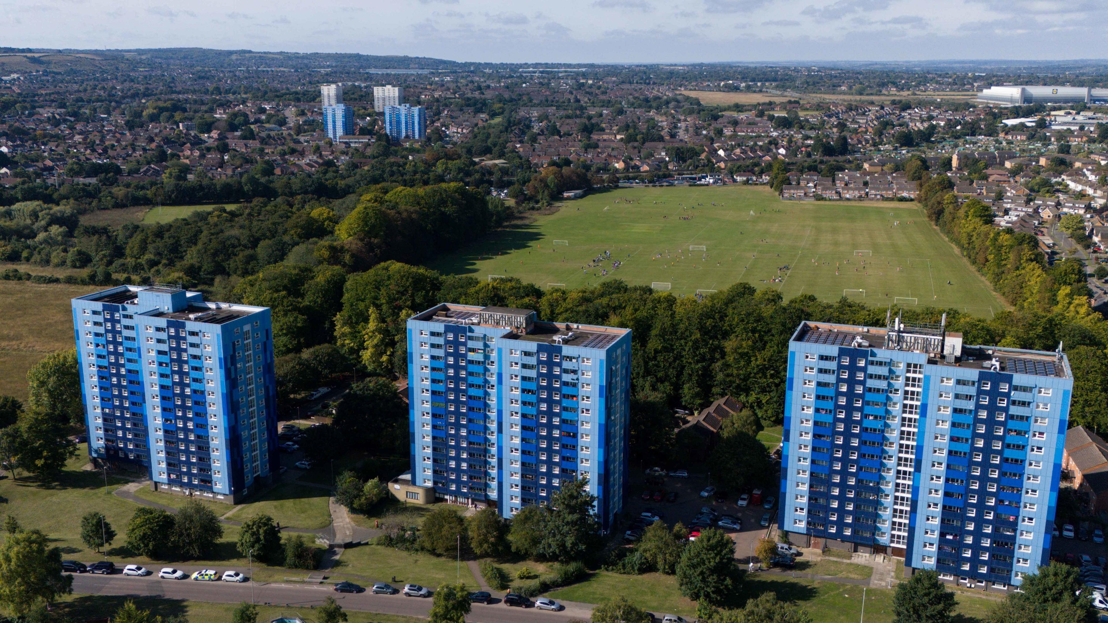 Aerial shot of three tower blocks, all in different shades of blue. Behind them is a large wooded area and an open expanse of playing fields, and more housing and tower bloc