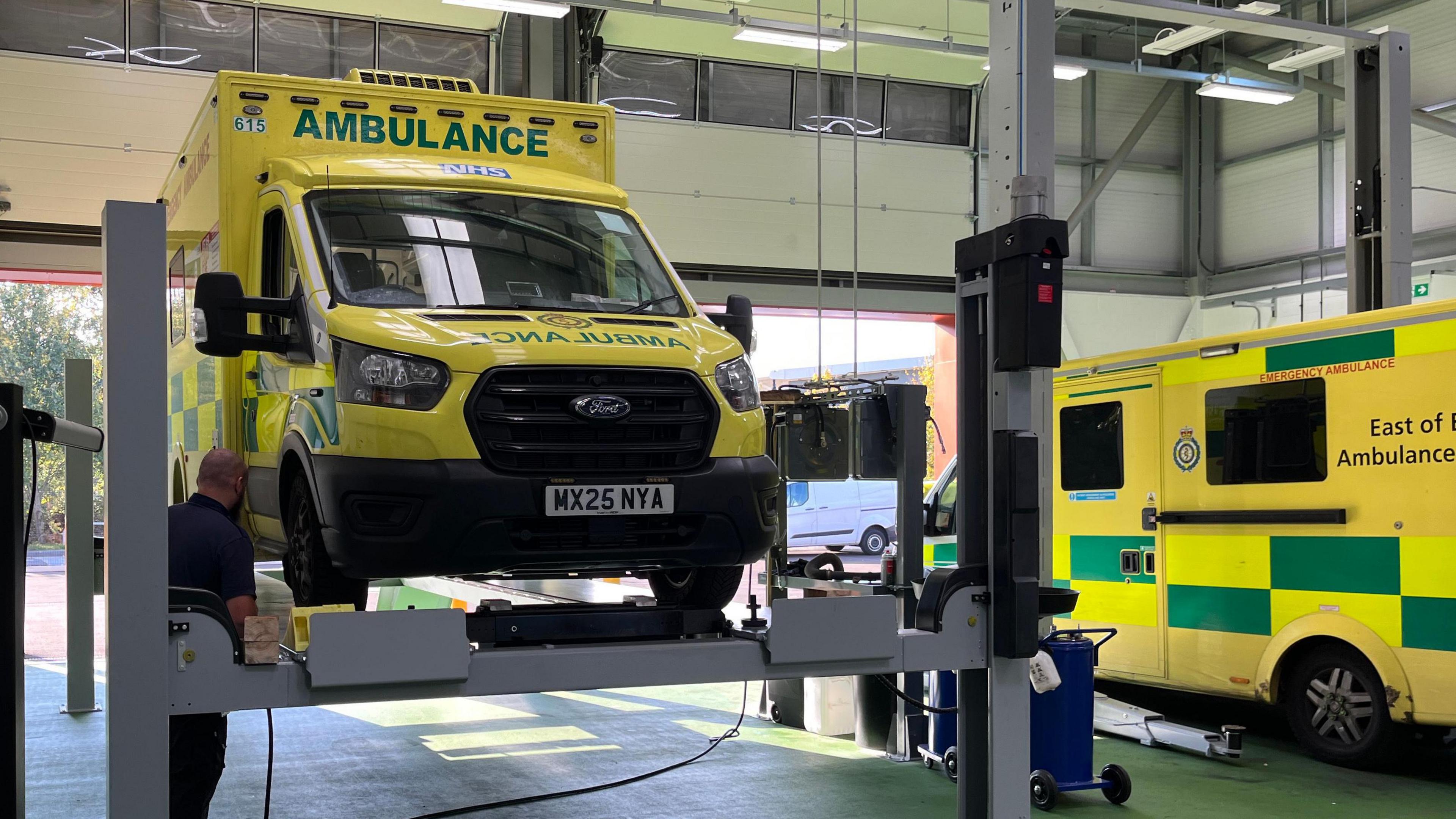 An ambulance, which was yellow and green and had AMBULANCE written on the front, was on a grey vehicle ramp in the middle of a room. Another ambulance is parked on the right.