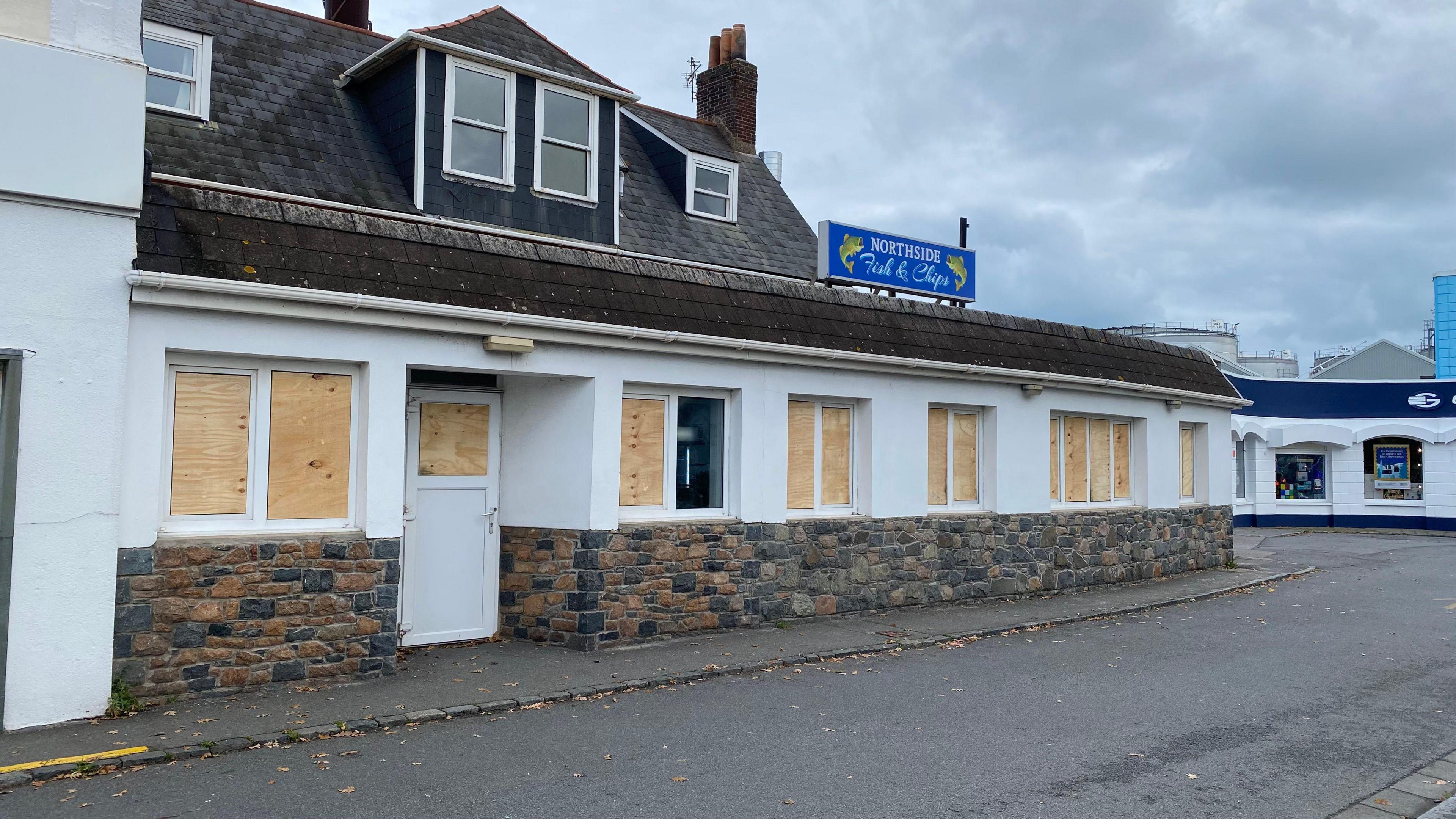 Boarded up windows at the front of Northside Fish and Chips shop. The building is white and has a small blue sign located on top at the centre. There is a brick wall at the front and a door to the left.