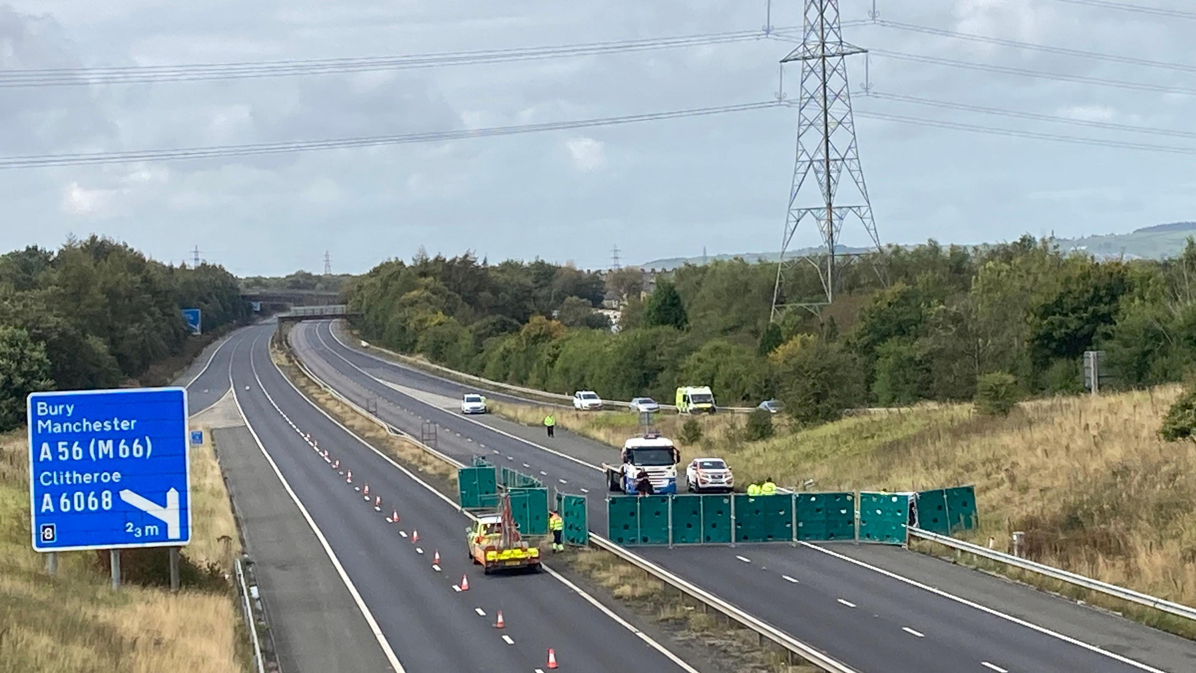 The motorway empty of traffic, with emergency service vehicles on the tarmac. A line of green panels marks the site of the accident. Cones are visible on the tarmac. 