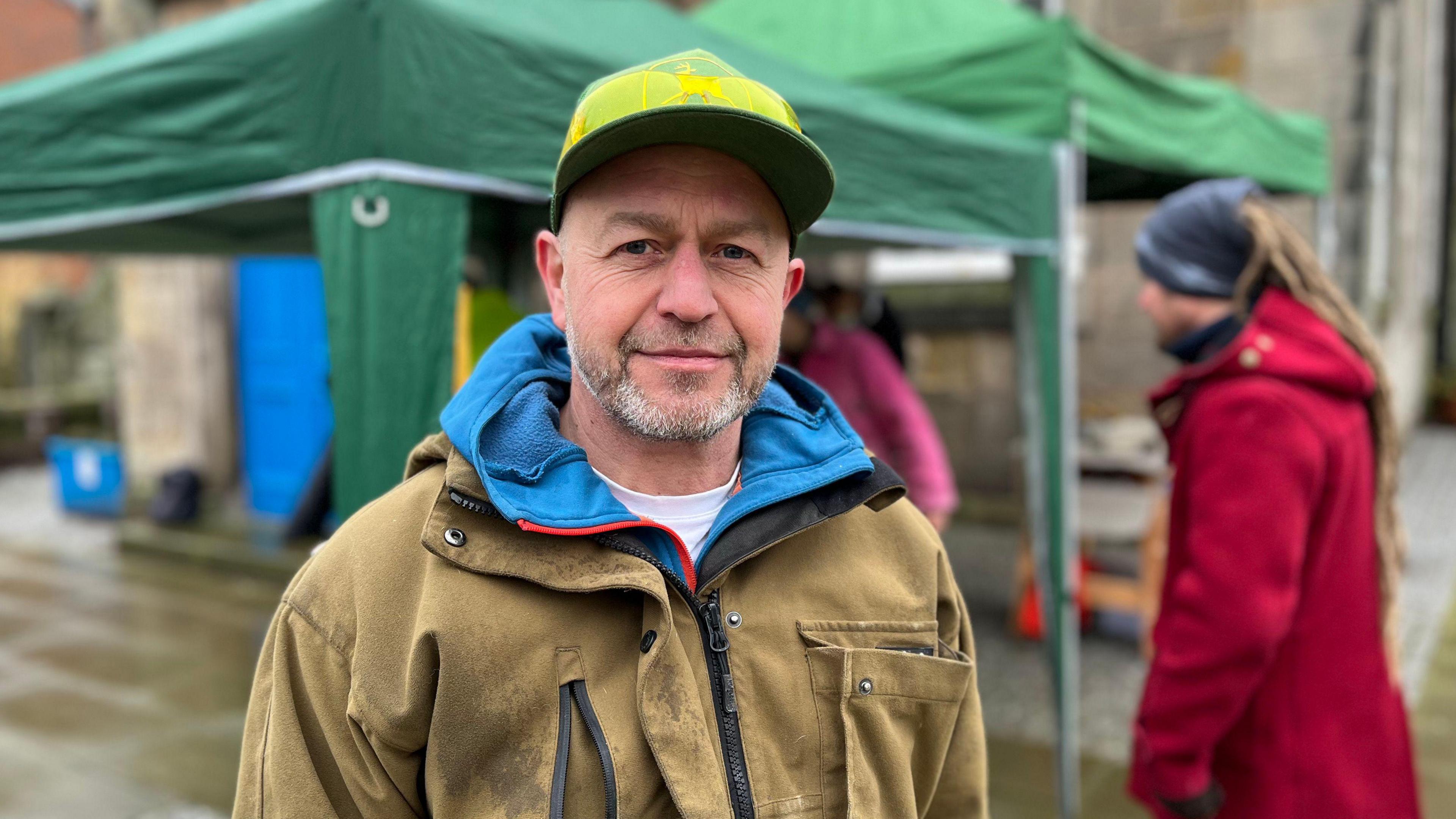 A man with grey beard, wearing a yellow cap and beige jacket, looks at the camera