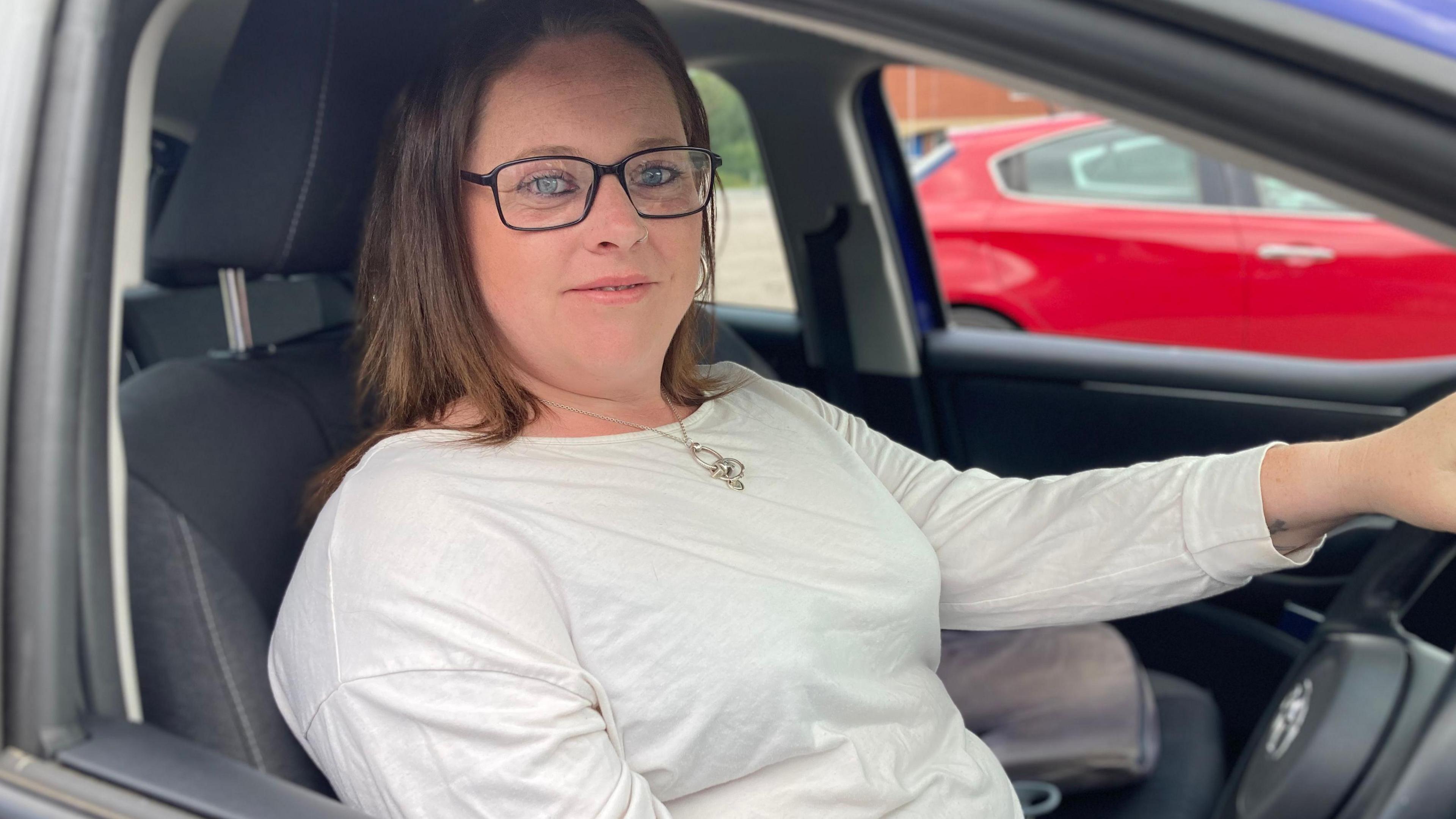 A bespectacled Lucy Bell sat in the driver's seat wearing a white top. She has brown shoulder-length hair.