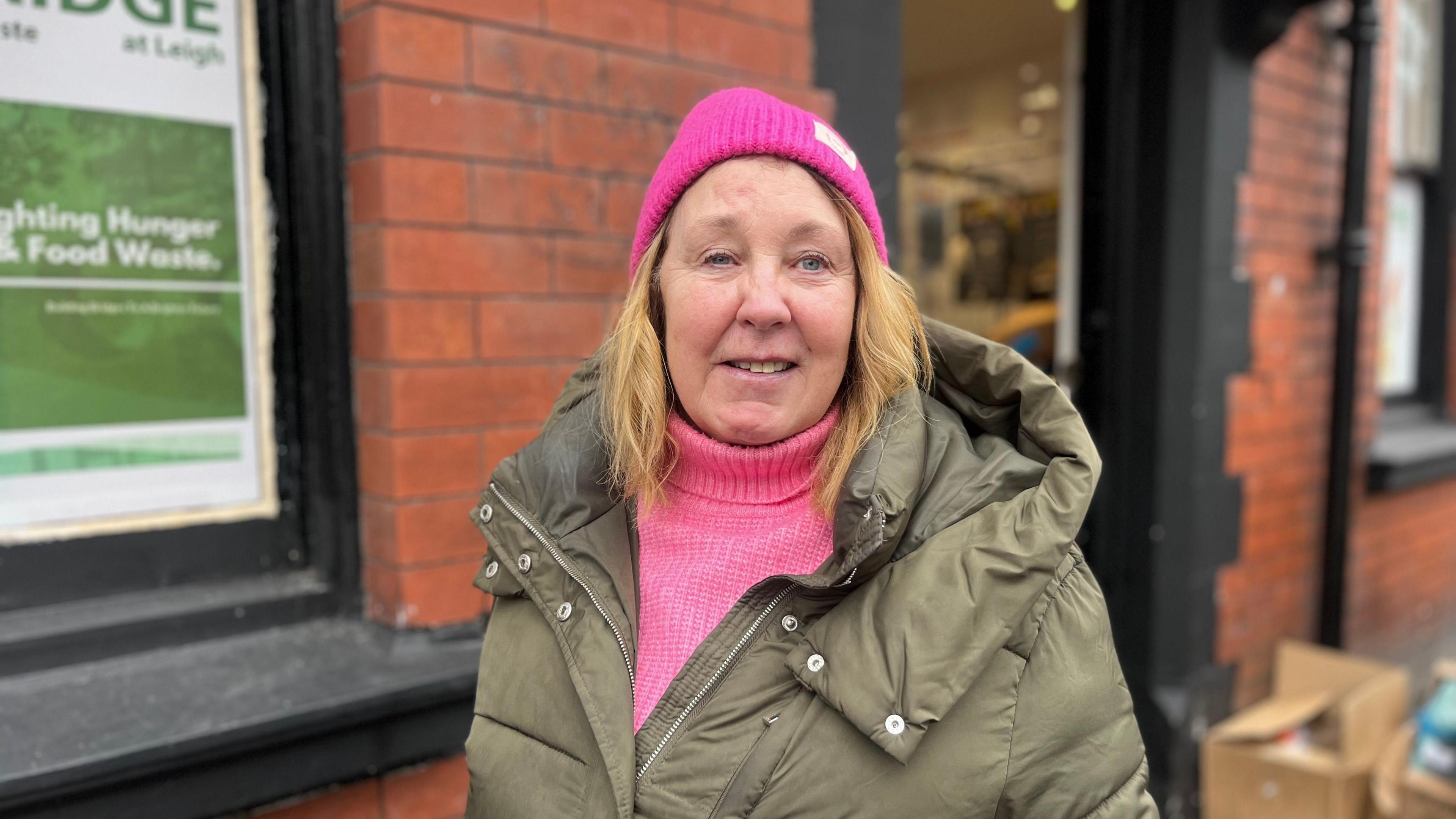 Ann Molineux is smiling as she stands outside the shop. She has shoulder length blonde hair and is wearing a pink woolly hat, pink roll neck jumper and a khaki green winter coat.