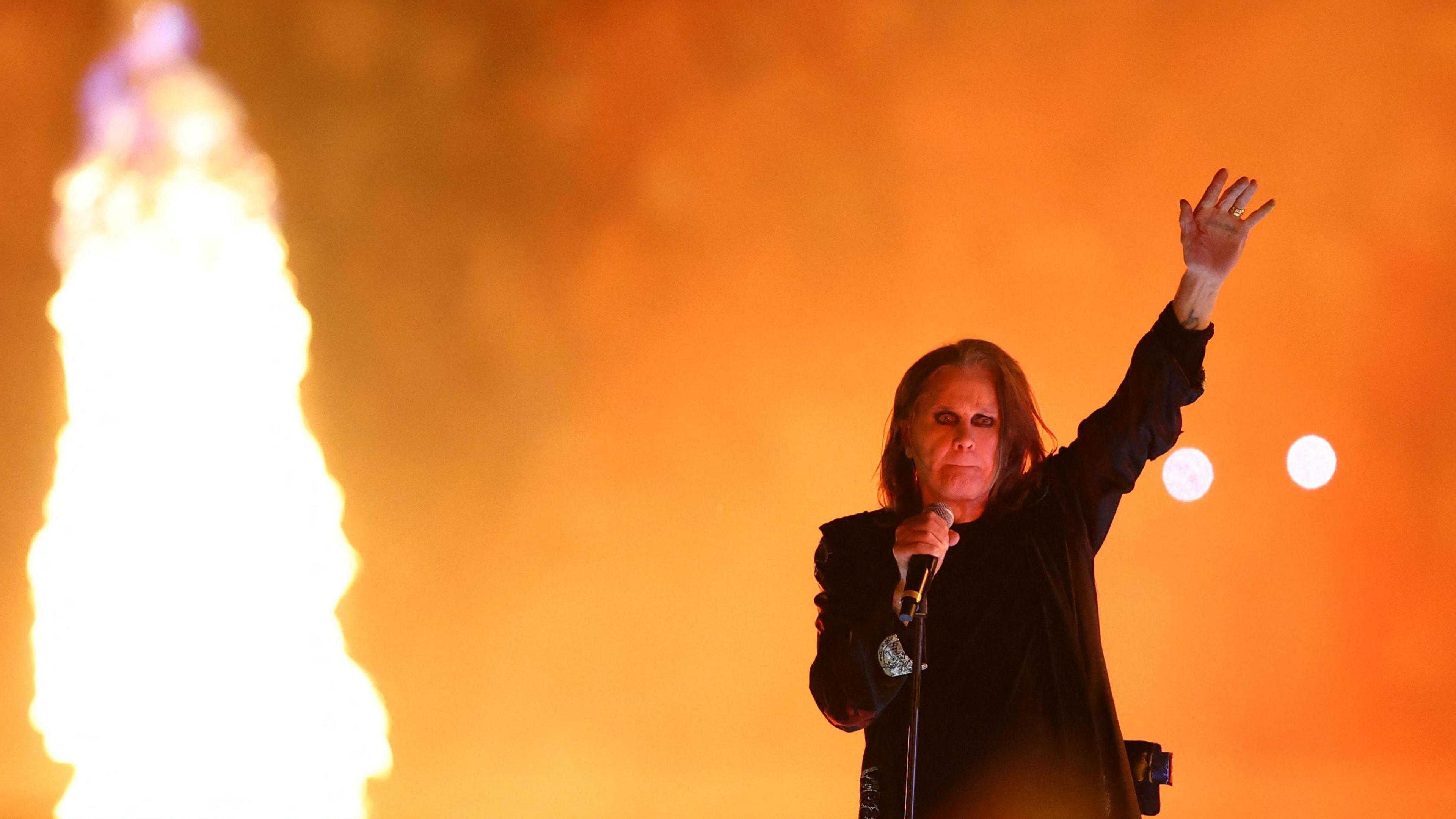 Image shows Ozzy Osbourne on stage in Birmingham at the closing ceremony of the Commonwealth Games. The background of the picture is orange and to the right is a large flame. Ozzy is on the left with his trademark long dark hair, black clothes and black eyeliner around his eyes. He is holding the microphone with his right hand and his left hand is in the air.