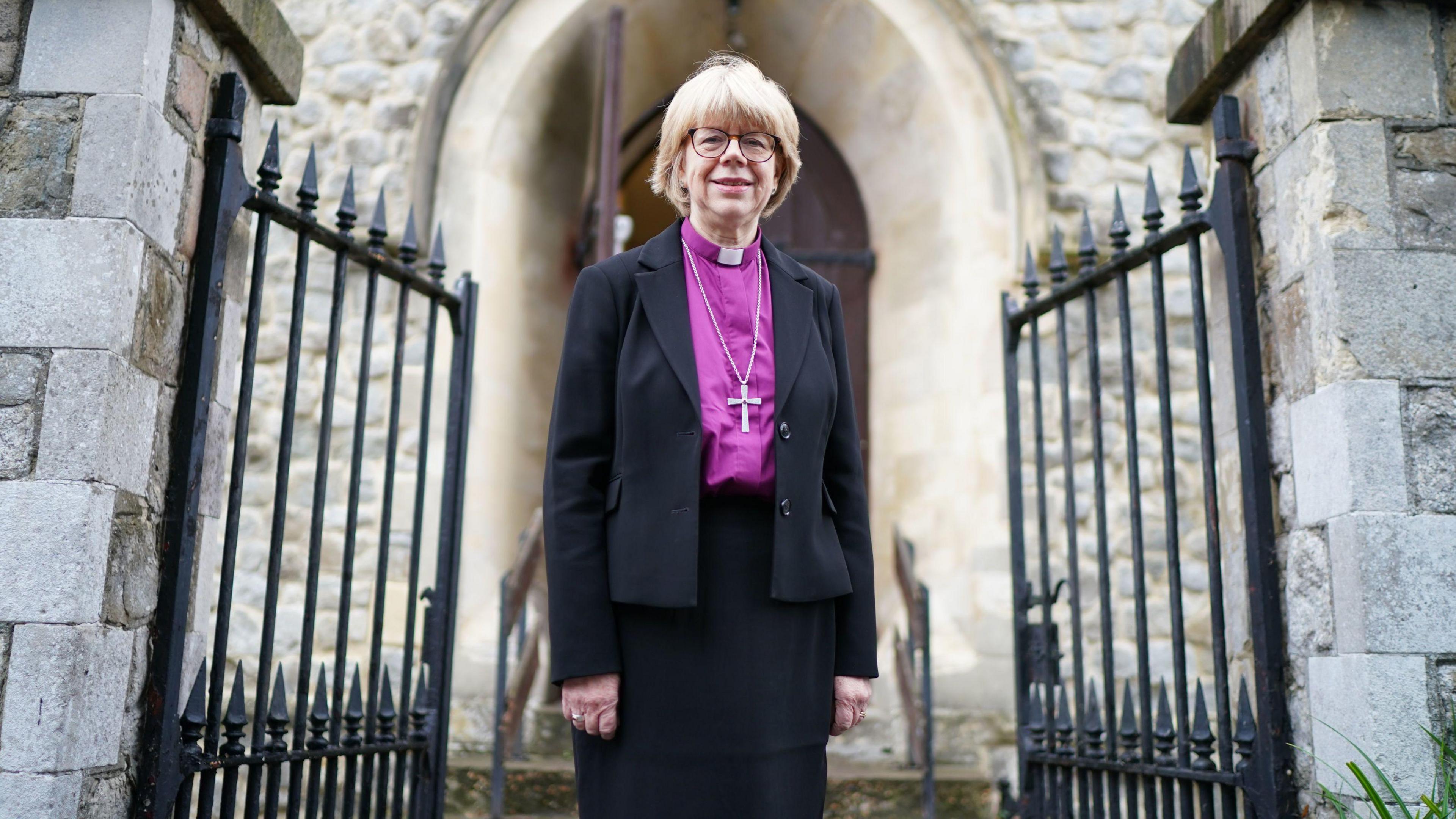 Dame Sarah Mullally stands in front of grey stone building in the middle of half opened black gates. She wears a black suit with a purple shirt, dog collar and cross around her neck.