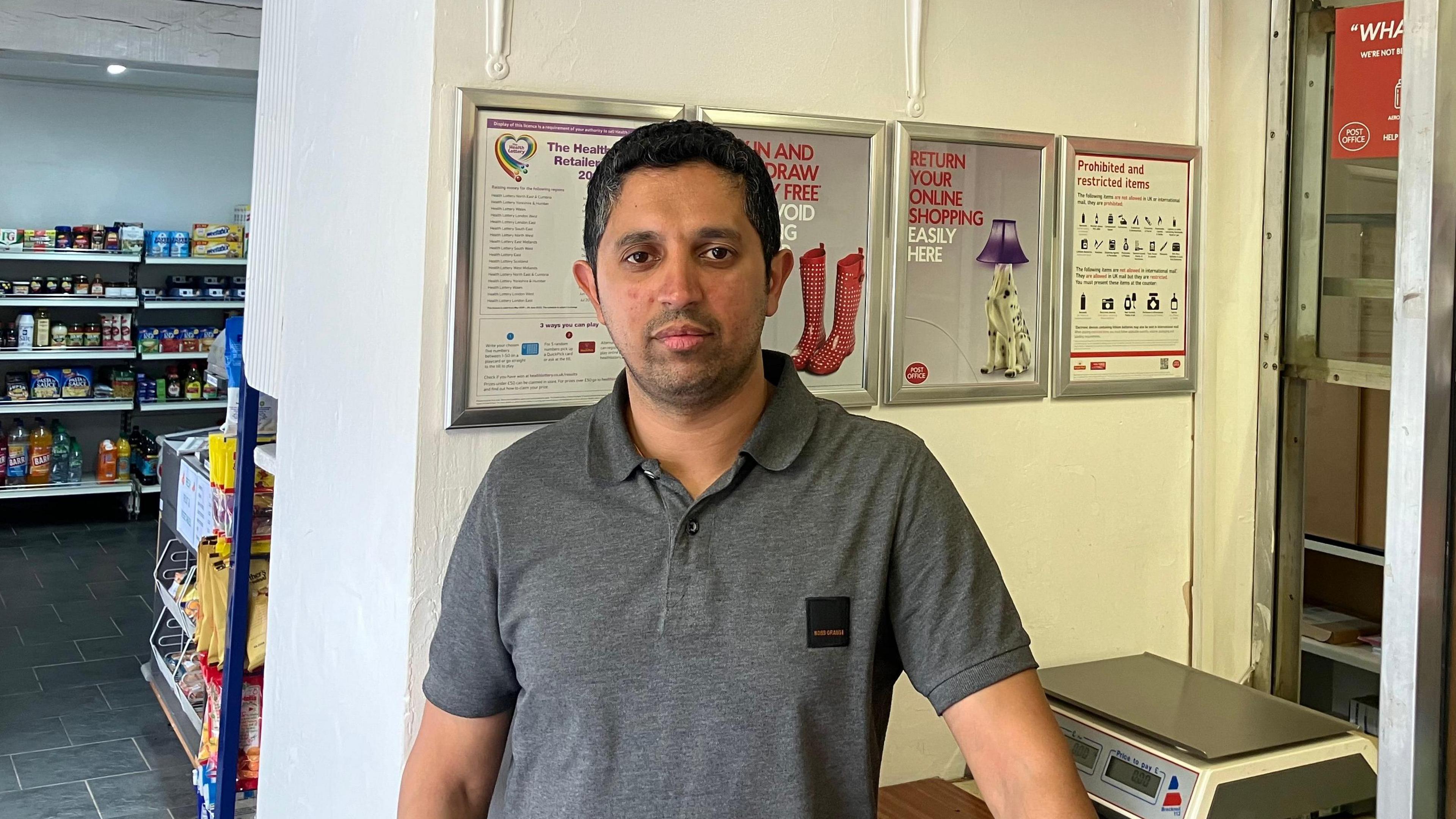 Mohammes Arif in a grey T-shirt standing in front of the postmaster's window in his shop