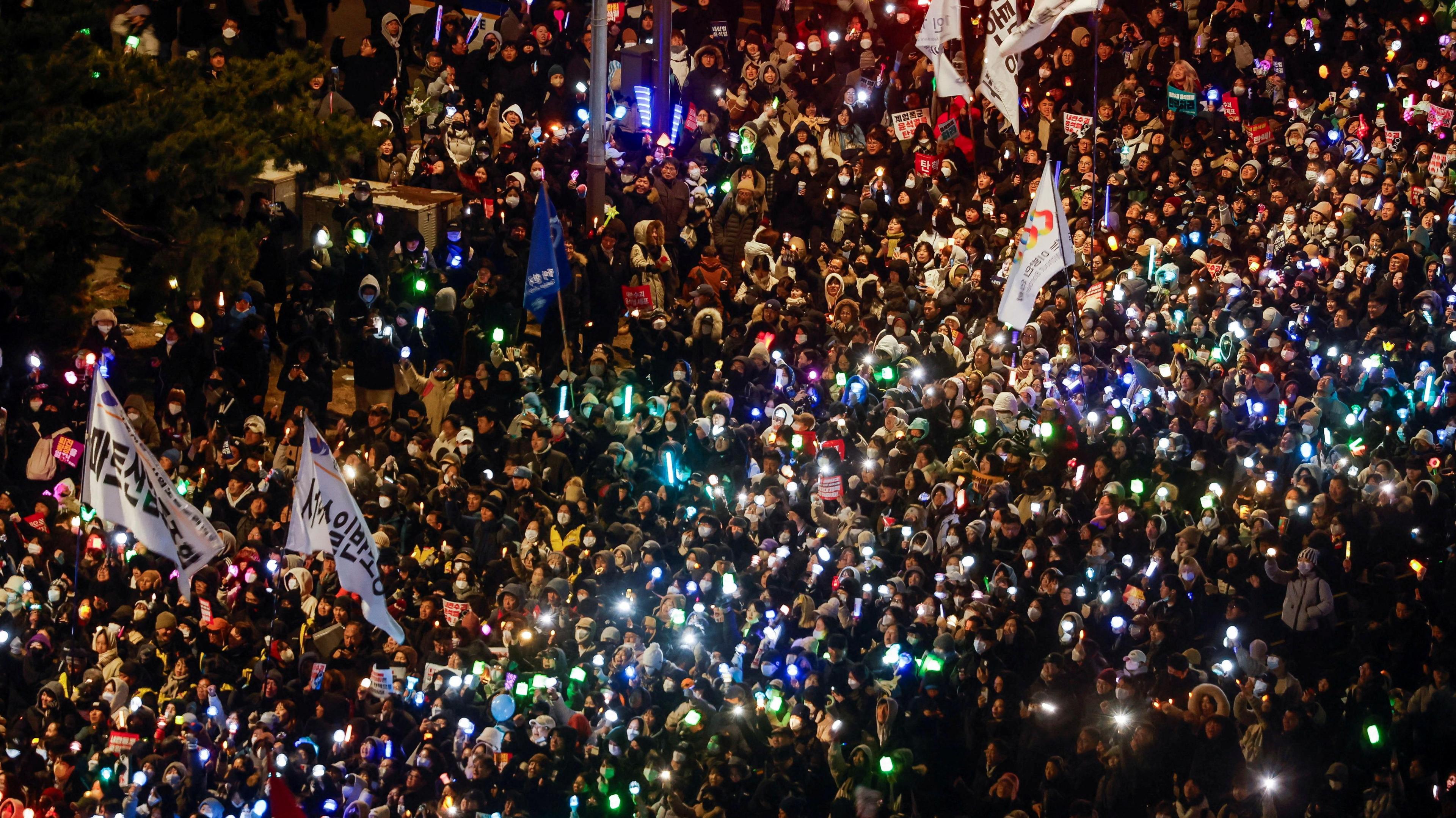 An aerial view of thousands of people at a protest in south Korea. There are several large banners waving above the crowd, and multiple lights twinkle in the dark.